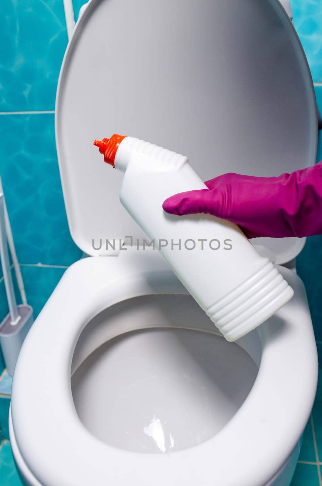 A hand wearing a pink glove carefully pours toilet cleaner from a bottle into the open toilet bowl, ensuring thorough cleaning.