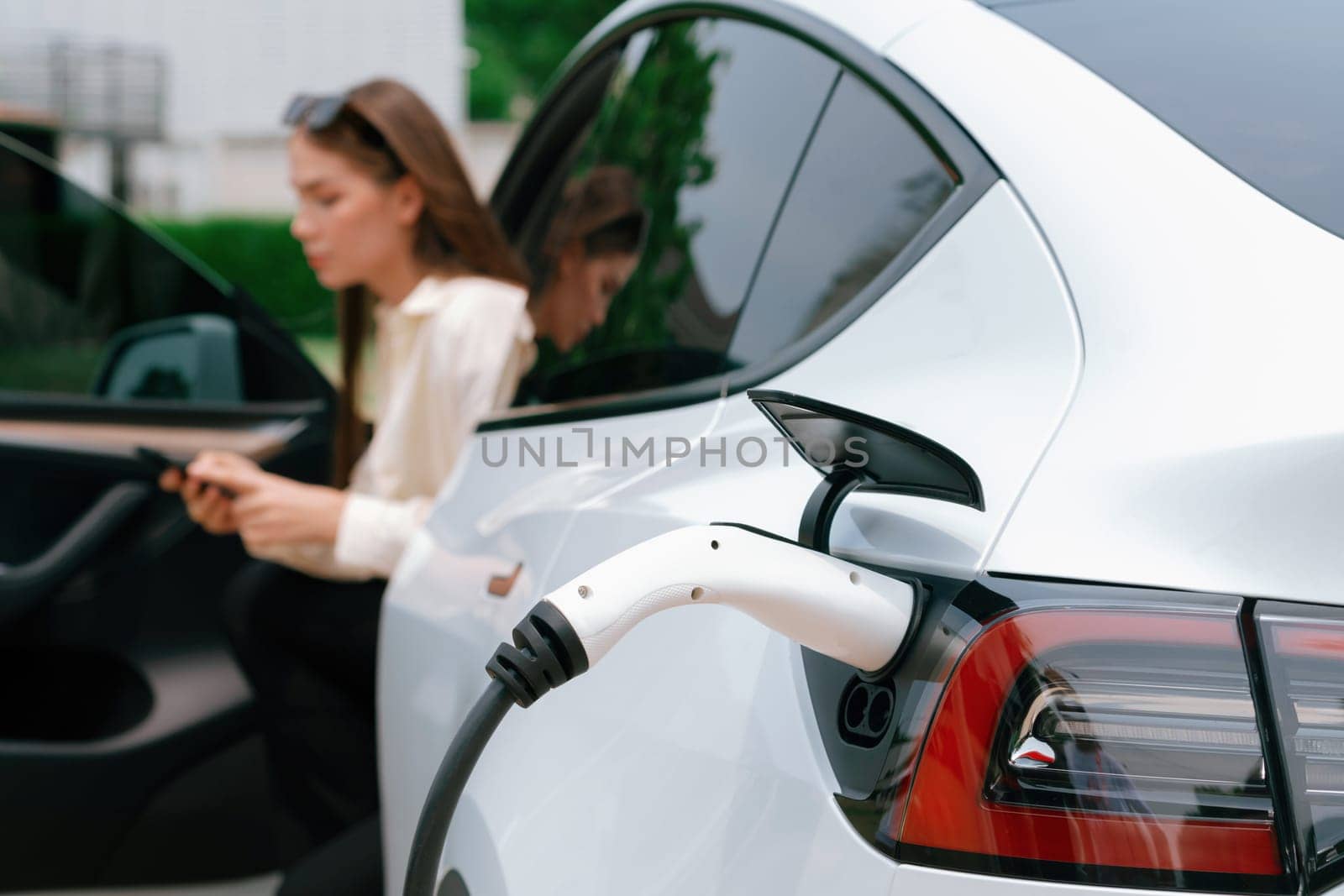 Young woman use smartphone to pay for electricity at public EV car charging station green city park. Modern environmental and sustainable urban lifestyle with EV vehicle. Expedient