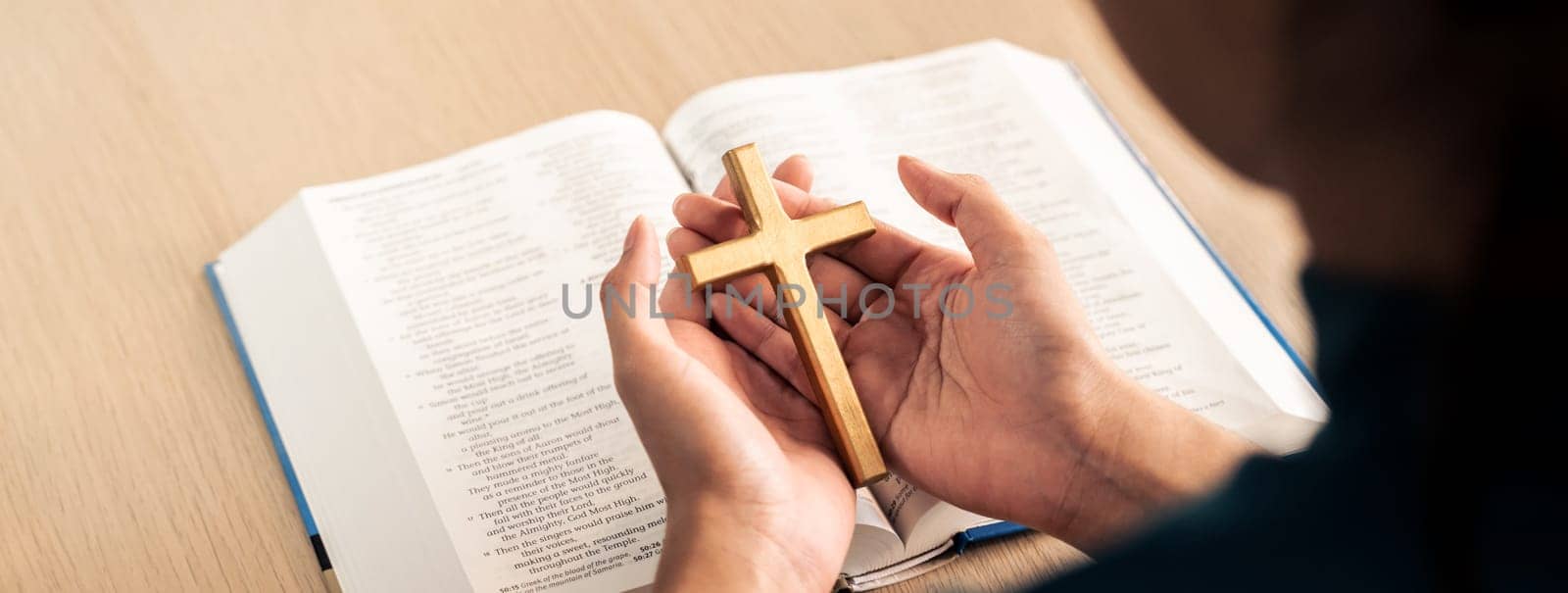 Female god believer holding wooden cross on opened holy bible book at light wooden church table. Top view. Concept of hope, religion, faith, christianity and god blessing. Warm background. Burgeoning.