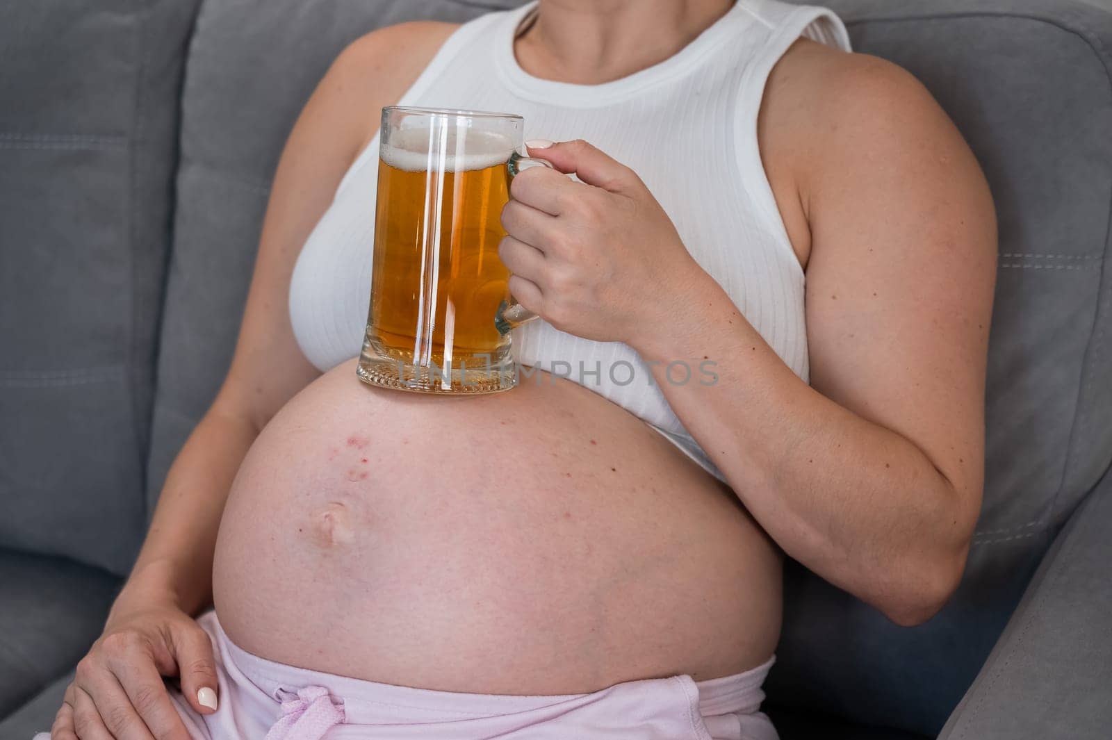 A faceless pregnant woman sits on the sofa and holds a glass of beer on her stomach. Skin rash. by mrwed54