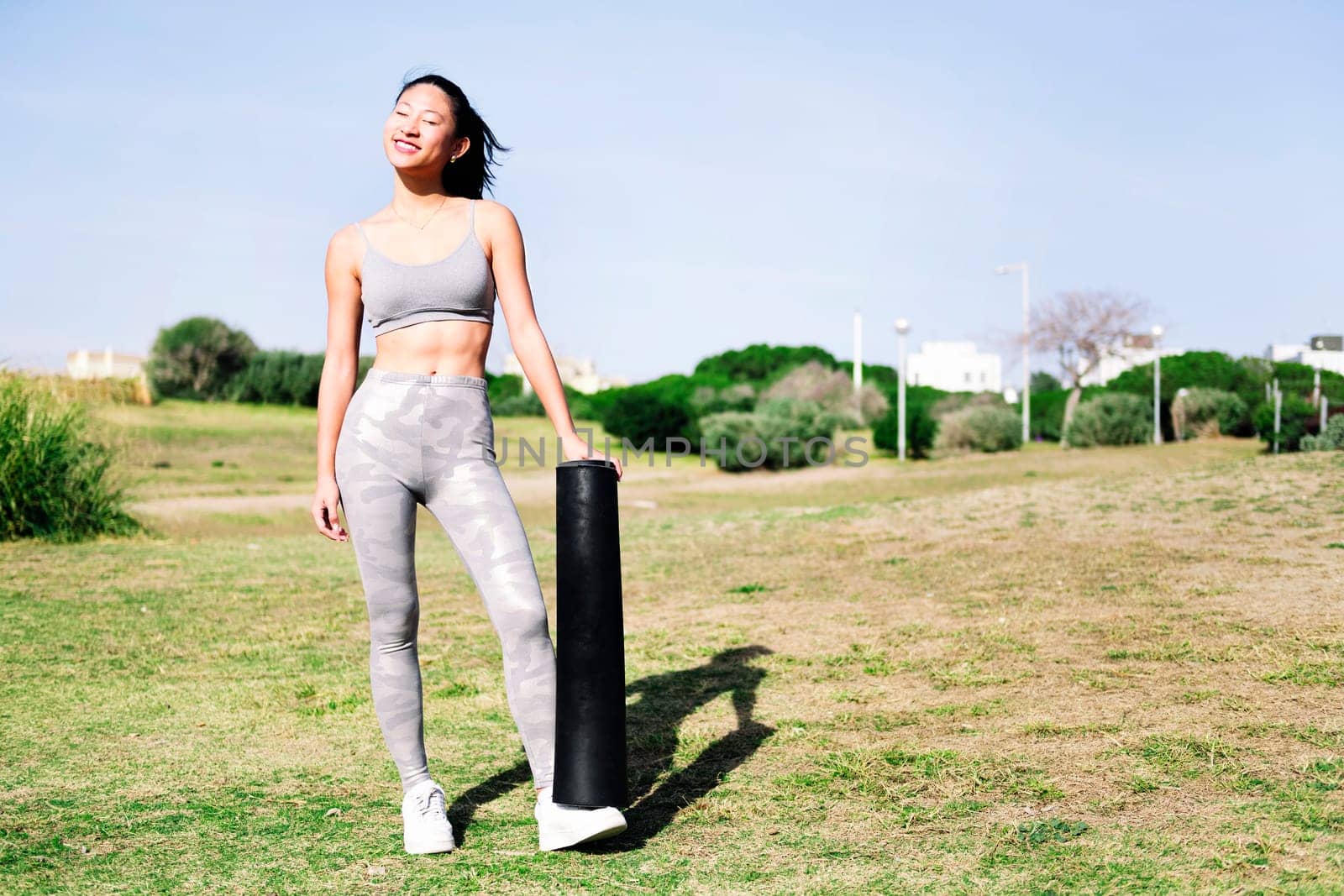 smiling young asian woman in sportswear ready with her mat for a yoga session on the grass in the park, healthy and active lifestyle concept, copy space for text