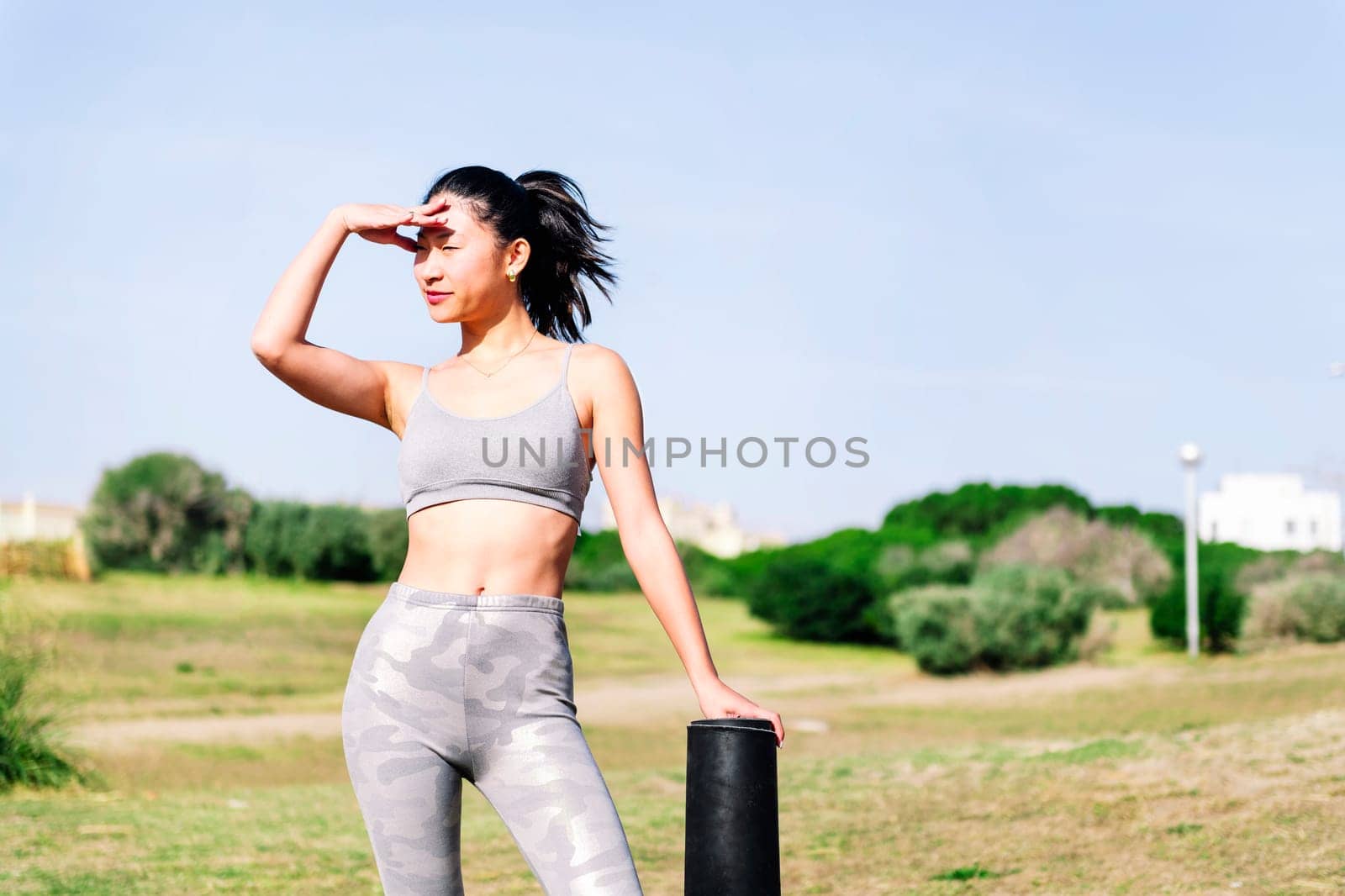 smiling young asian woman in sportswear ready with her mat for a yoga session on the grass in the park, healthy and active lifestyle concept, copy space for text