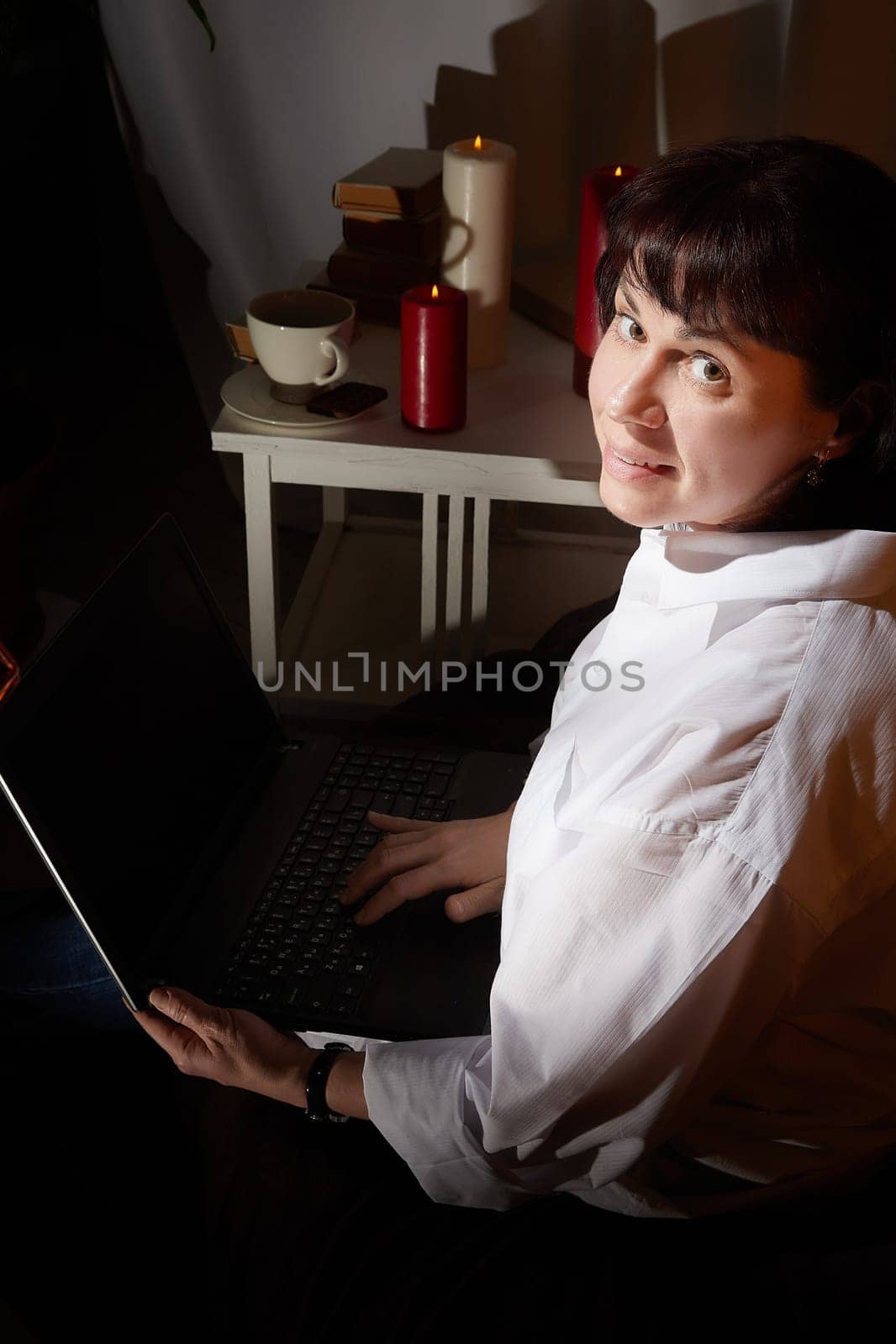 A girl in white shirt with a laptop in a dark room. A middle-aged woman works online remotely in the evening. The student is preparing for the exam at night. Blogger or freelancer in the chat