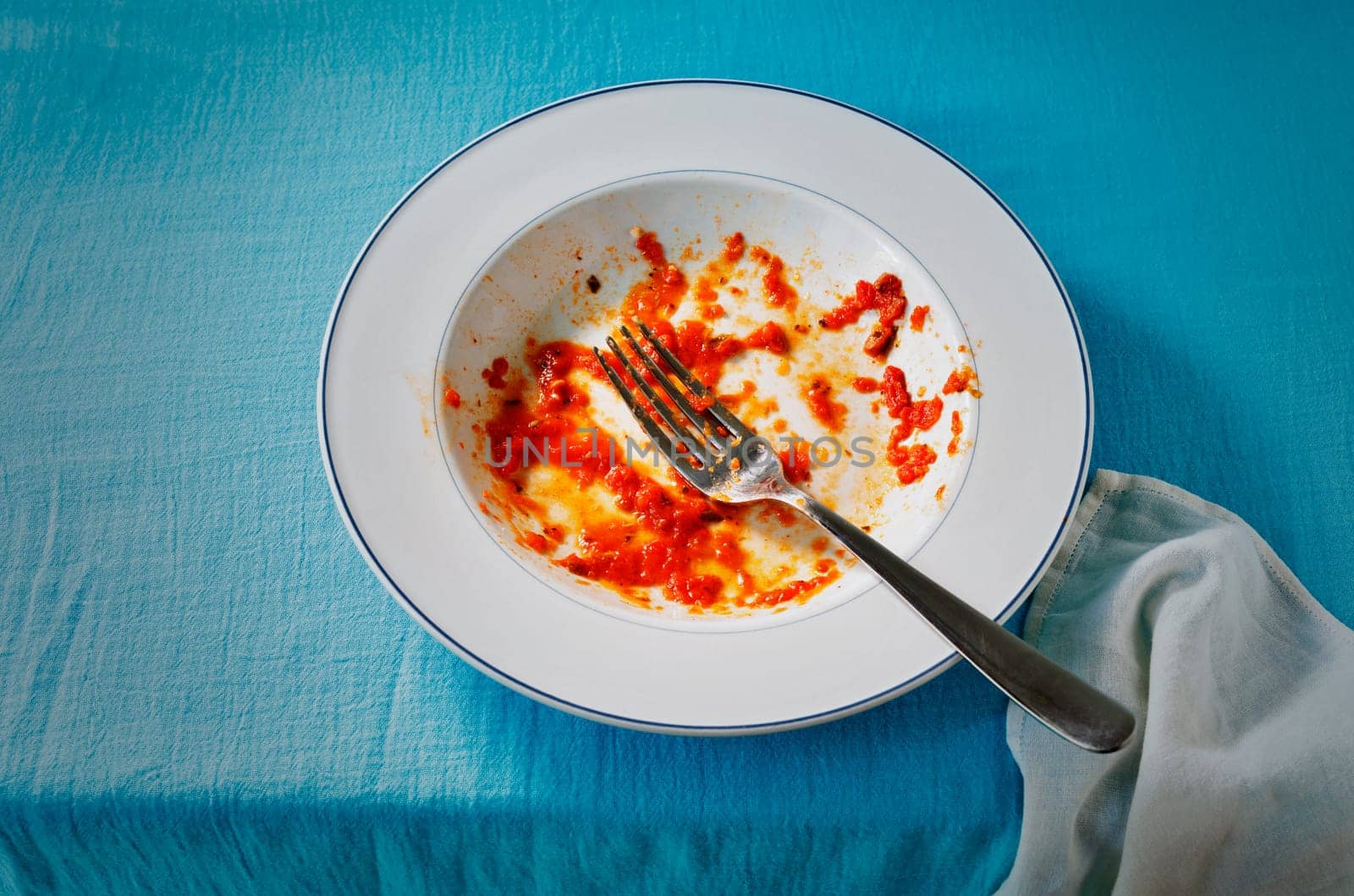 White dirty plate with red tomato sauce leftover and fork on table with blue cloth