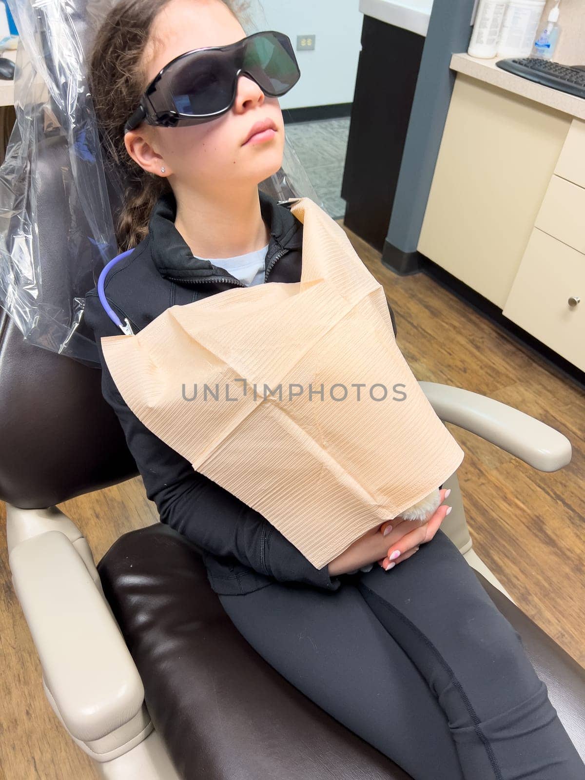 In the dentist chair, a young girl sits patiently, clad in protective eyewear and a paper bib, awaiting a dental treatment with calm and composure.