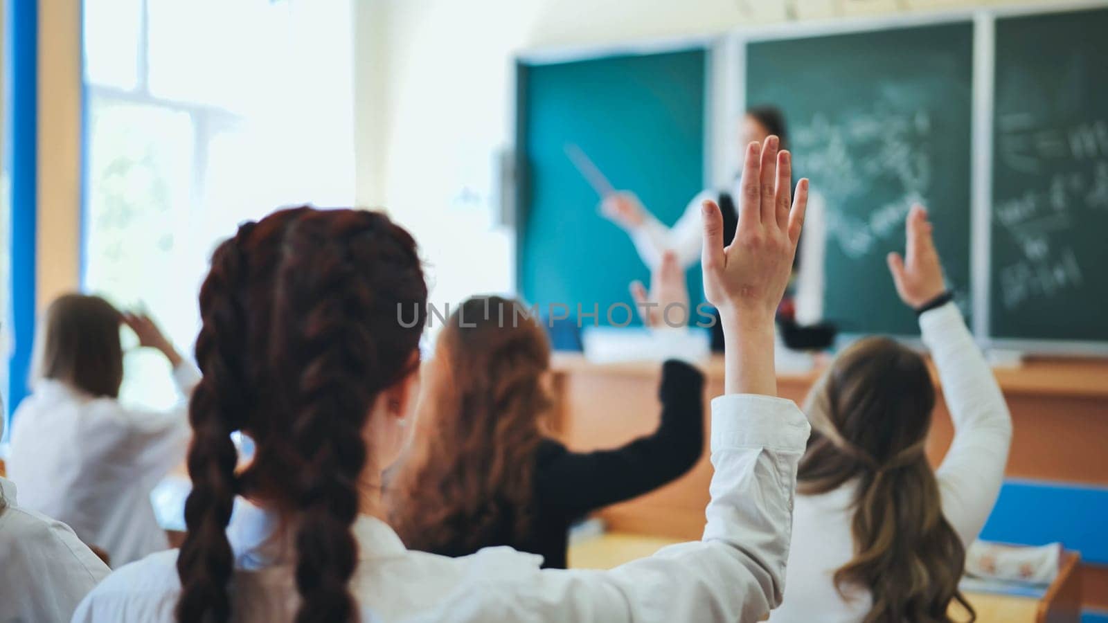 Intelligent group of young school children all raising their hands in the air to answer a question posed by the female teacher, view from behind by DovidPro