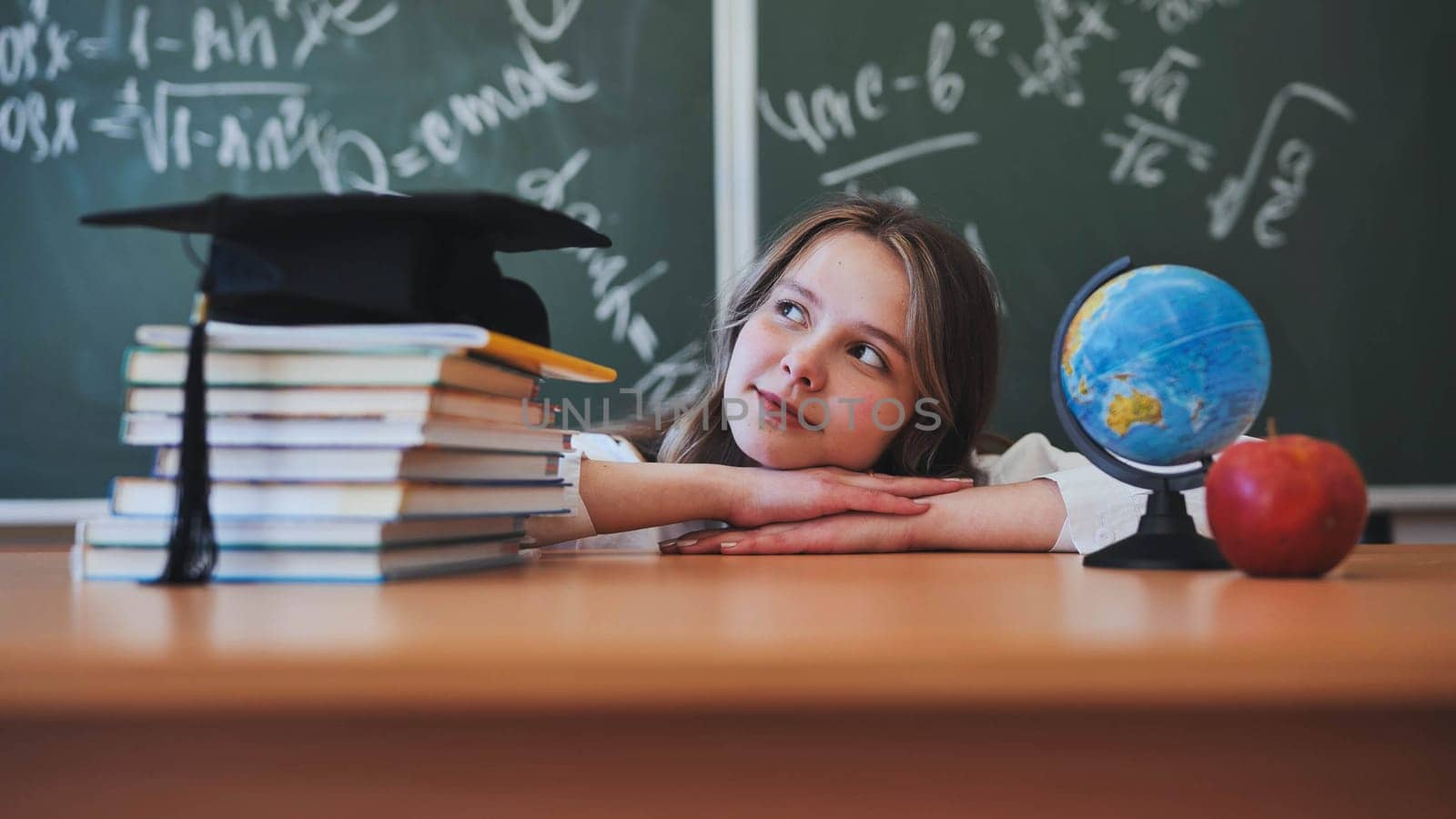 Adorable school girl posing at her desk against a background of blackboard, books, globe and graduation cap. by DovidPro