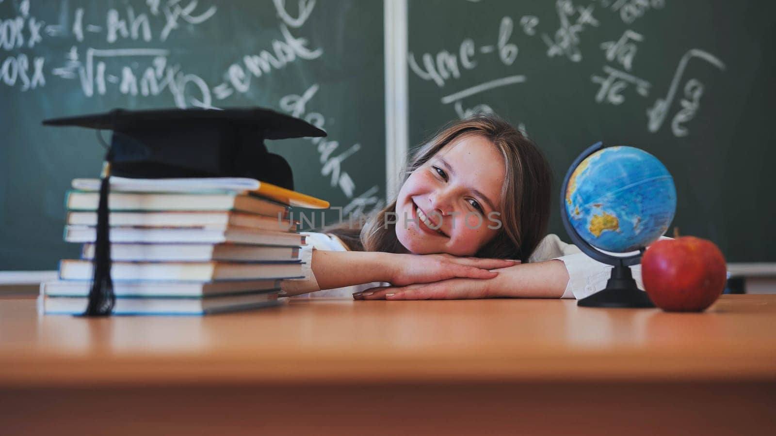 Adorable school girl posing at her desk against a background of blackboard, books, globe and graduation cap. by DovidPro