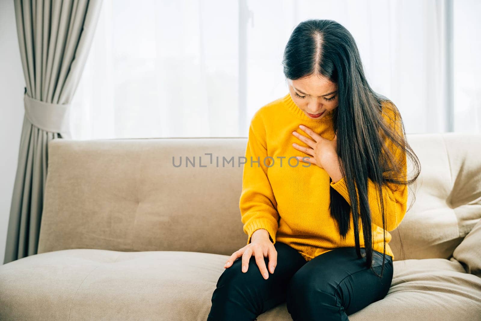 Portraying a heart attack, Asian woman on sofa holds her left chest in pain requiring immediate medical assistance. Illustrating heartache discomfort and the need for emergency aid.