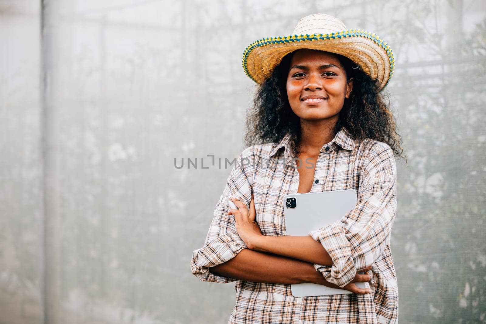 PhotoSuccessful greenhouse owner a young woman in a checked shirt and apron stands with arms crossed smiling of a woman posing confidently with a straw hat and crossed arms by Sorapop