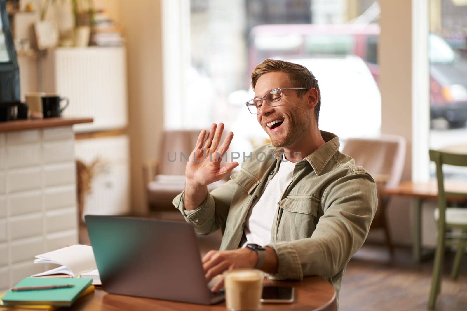 Portrait of handsome, smiling young man, online tutor, businessman working in cafe remotely, waving hand at laptop, connects to video chat or meeting.