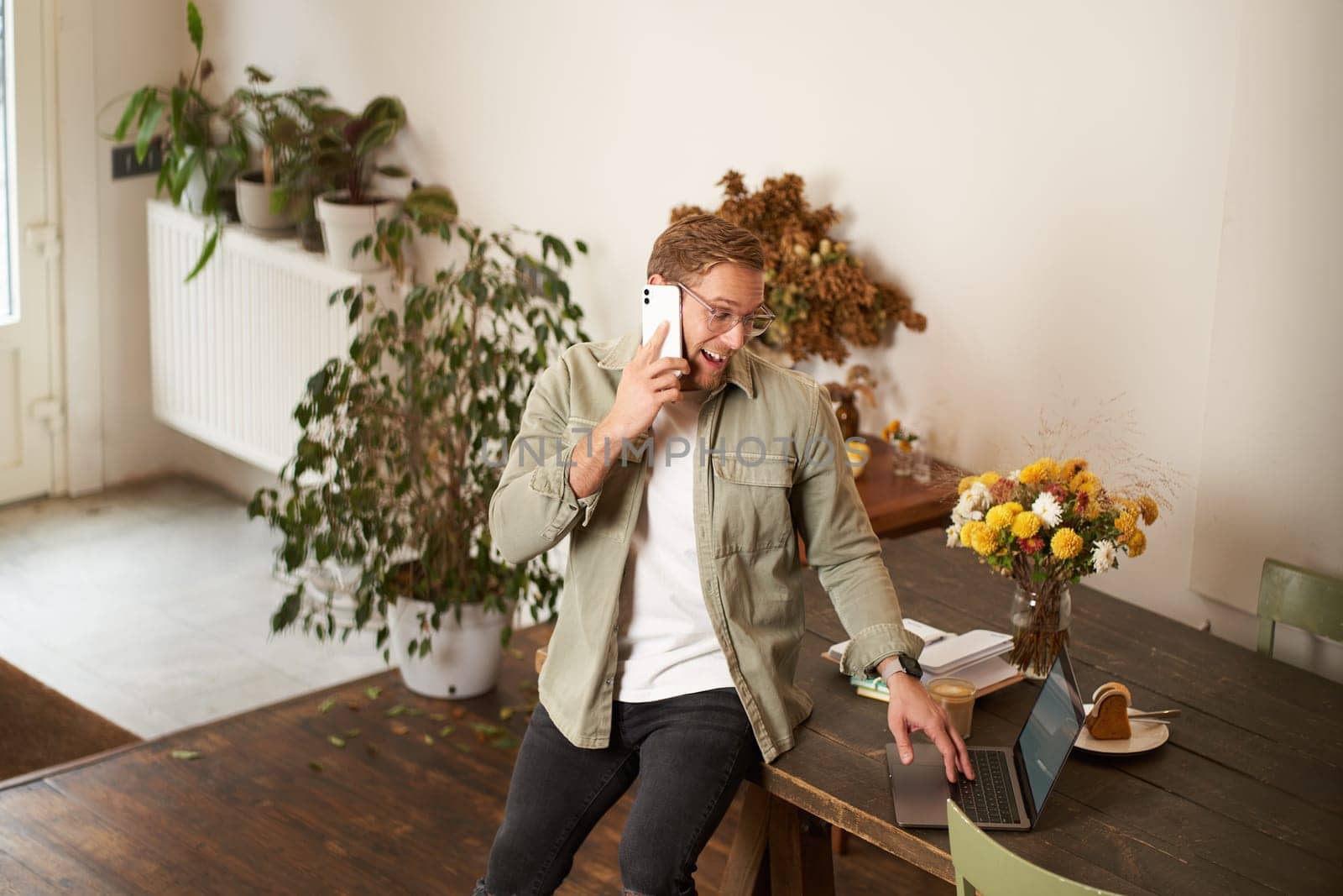 Portrait of happy young manager, man having negotiations, business conversation over the phone, talking on mobile, sitting in office on table with laptop.