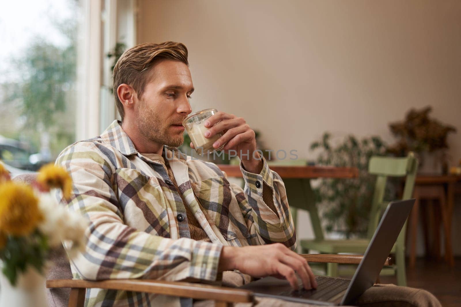 Portrait of handsome young man, working in cafe, sitting with laptop and coffee in hand, has remote job and using local co-working space instead of going to the office.