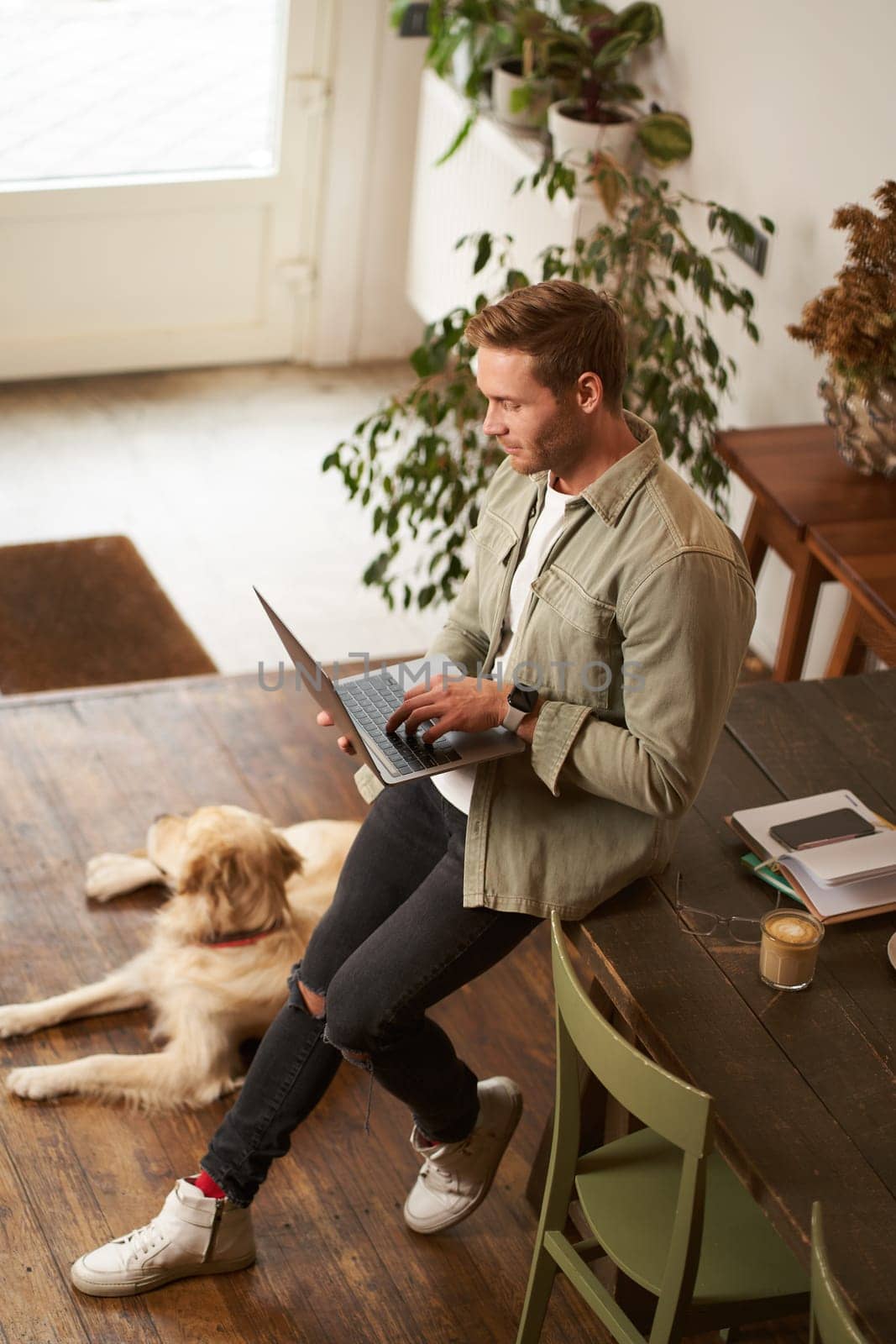 Vertical shot of handsome businessman, young man with laptop leans on table, sitting at co-working space with his dog. Young salesman checks his messages, smiling, working in an office with a pet.