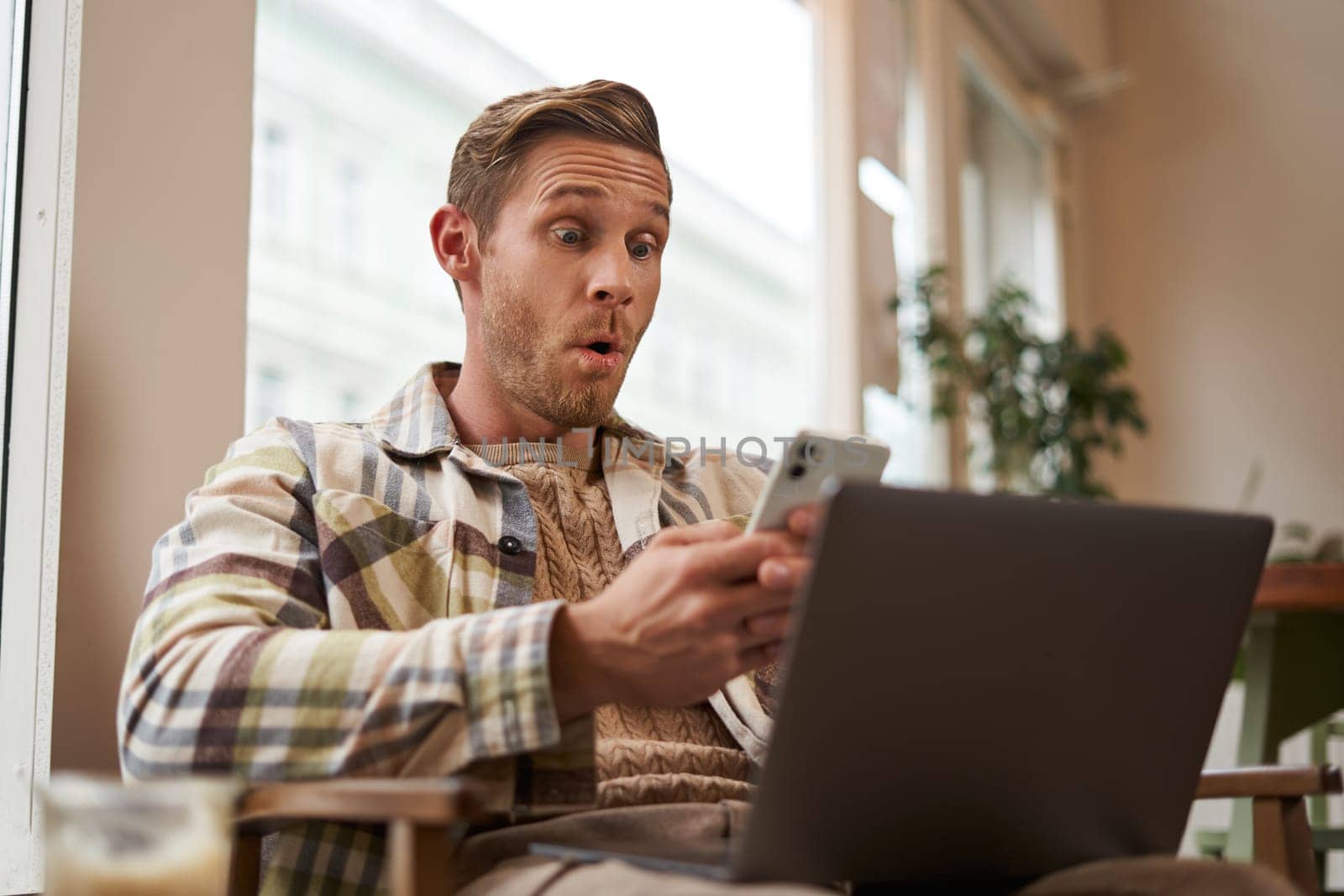 Image of man in cafe, coffee shop visitor sitting in chair with laptop and smartphone, looking surprised and excited at mobile phone screen, amazed by big news, promo offer on app.
