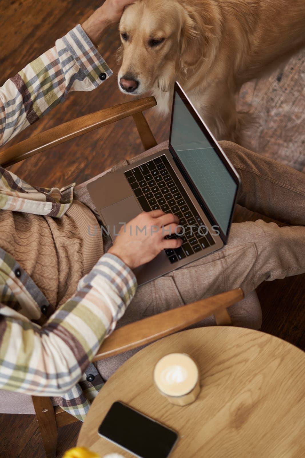 Vertical cropped photo of male hands and body, man petting the dog while working on laptop in cafe, drinking coffee.