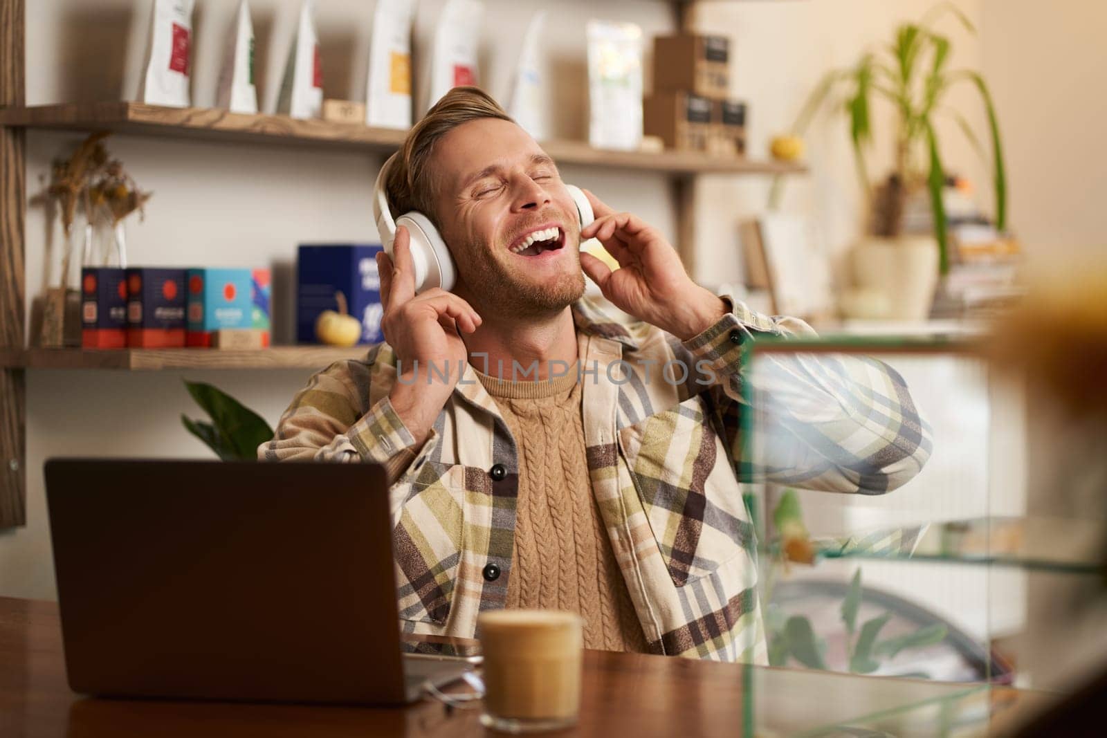 Lifestyle portrait of happy, excited young man, sitting in coffee shop with laptop and headphones, dancing on his chair and enjoying great quality sound, streaming favourite songs.