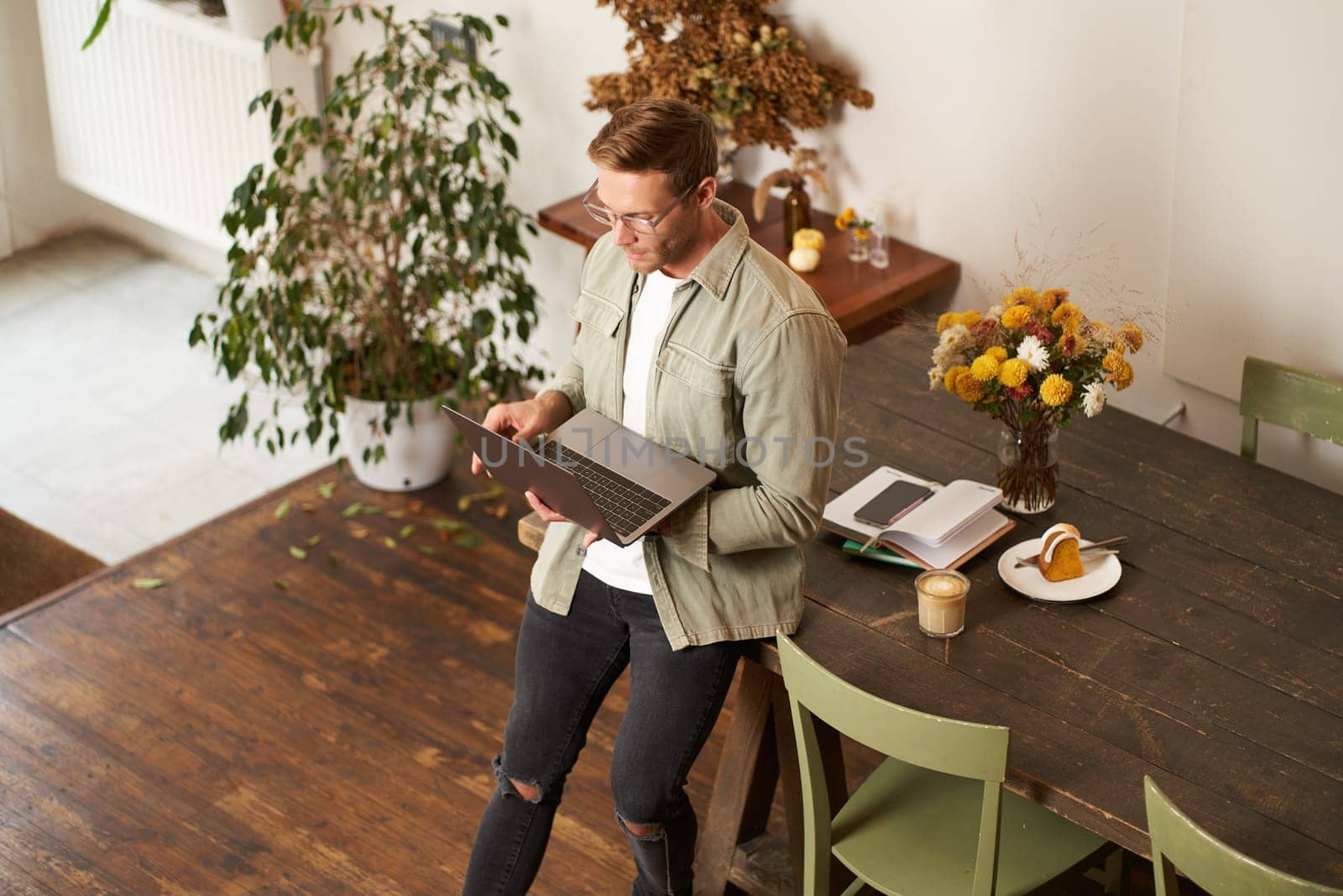 Image of young man, successful businessman sitting on table, looking at laptop, working in an office, looking concentrated on the project, doing job task, drinking coffee.