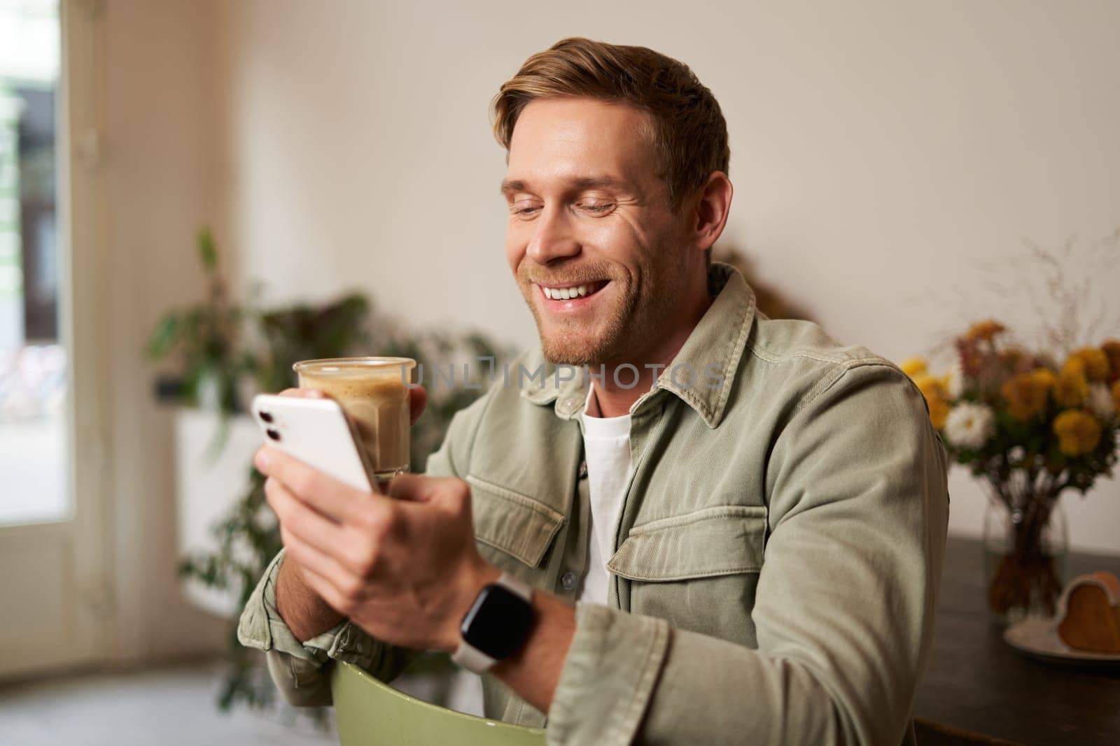 Portrait of smiling charismatic young man, sitting in coffee shop, drinking cappuccino, looking at his mobile phone, reading a message by Benzoix