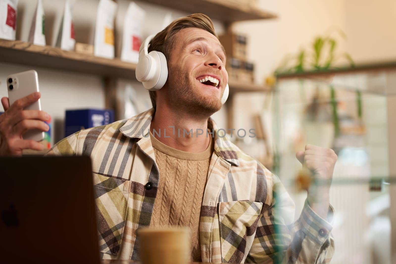 Lifestyle portrait of happy, excited young man, sitting in coffee shop with laptop and headphones, dancing on his chair and enjoying great quality sound, streaming favourite songs.