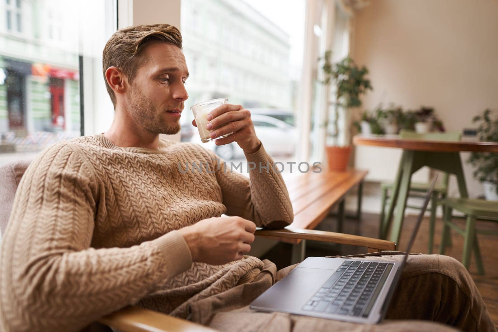 Working people and public space concept. Young handsome man, ux ui designer sitting with laptop in cafe, drinking cappuccino, looking at screen with serious face by Benzoix