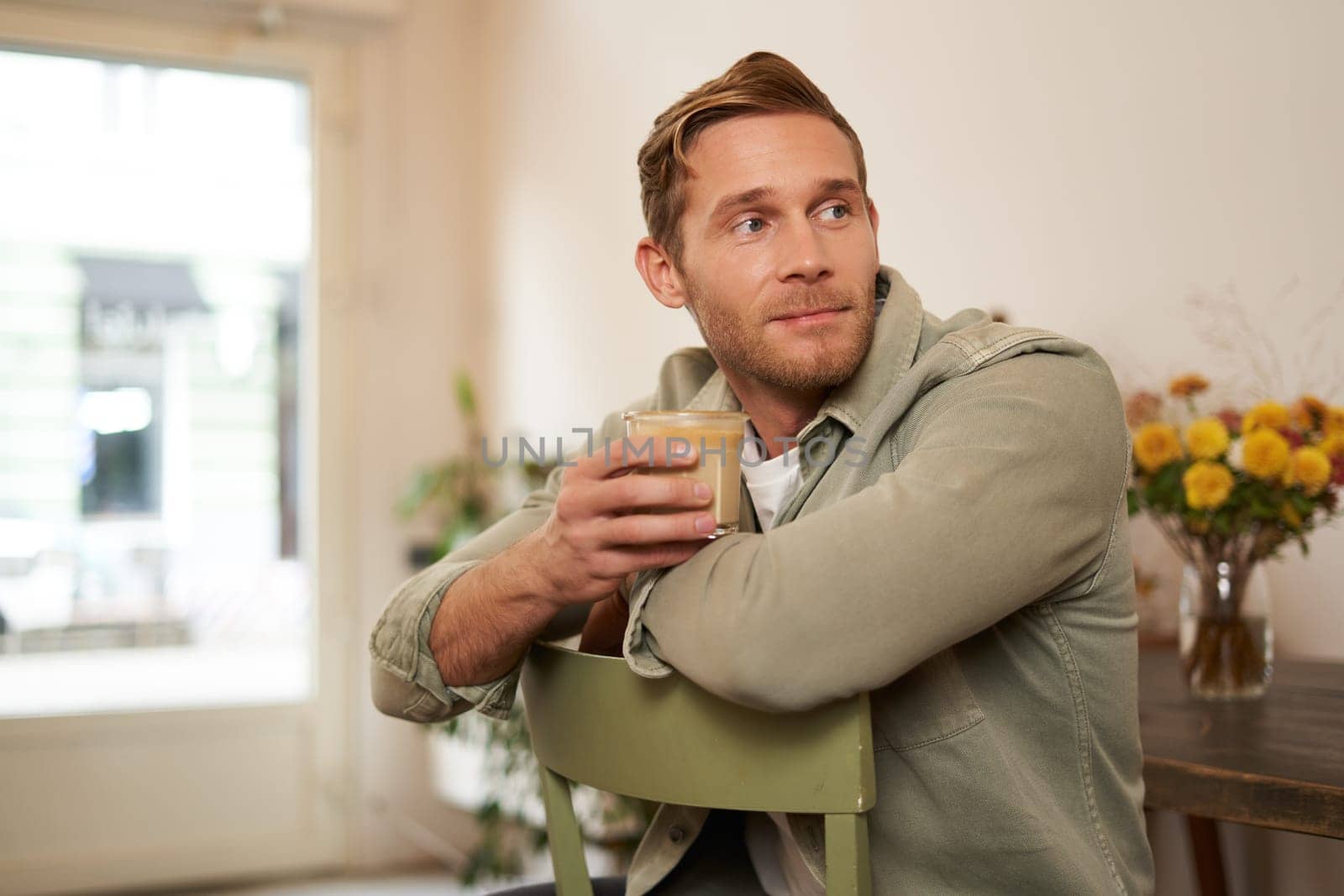 Portrait of good-looking young man with cup of coffee, sitting on chair in cafe, smiling and relaxing with his cappuccino drink by Benzoix