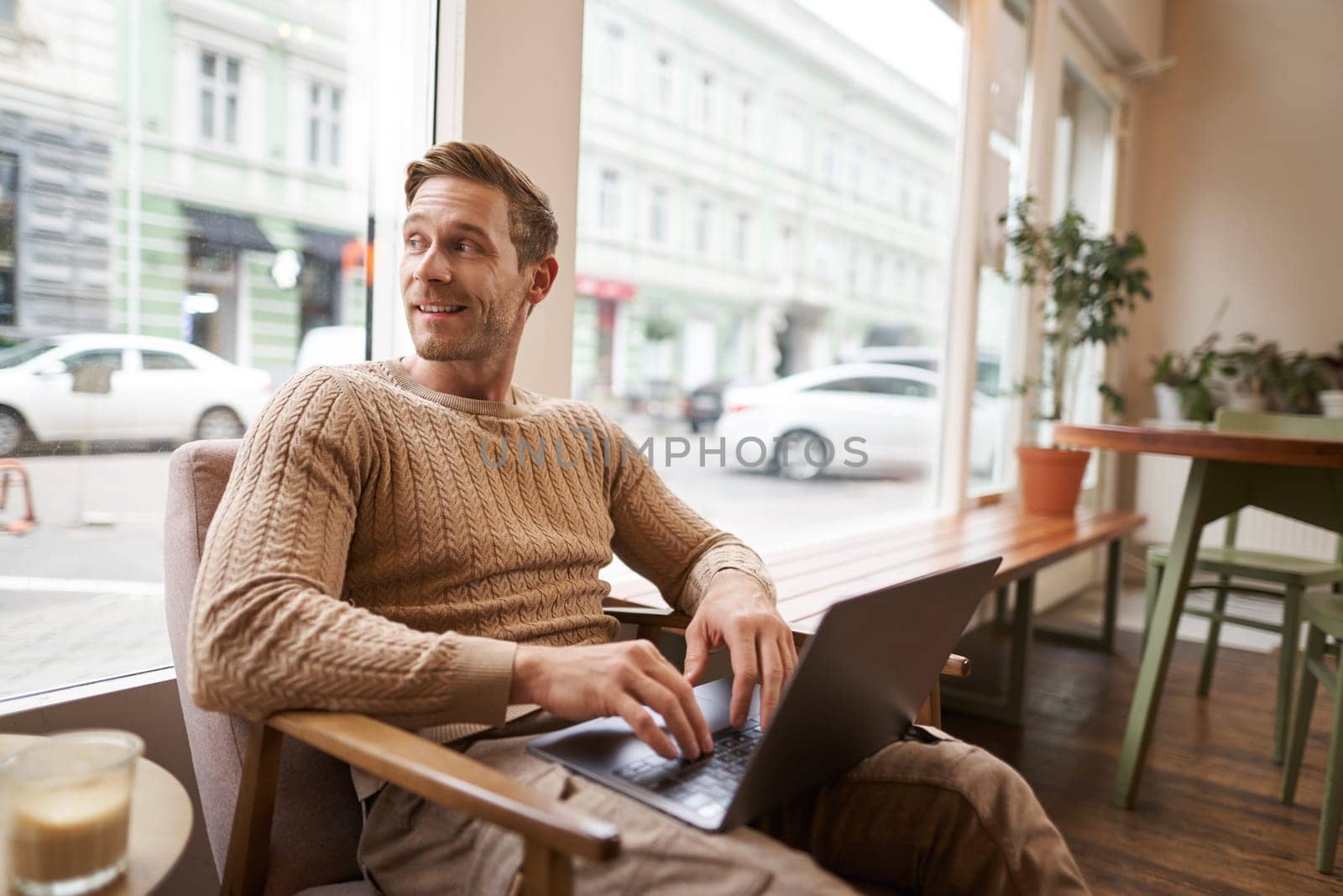 Portrait of young handsome businessman, a man with laptop, sitting in coffee shop on chair and working, freelancer doing his job online from cafe.