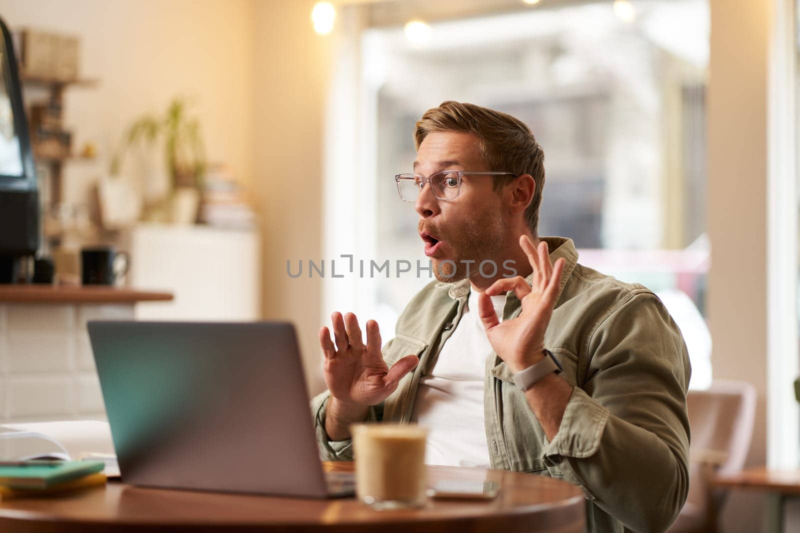 Portrait of man giving lessons online, shows okay, ok hand sign at laptop camera, talking to someone, having a meeting video chat, sitting in cafe.