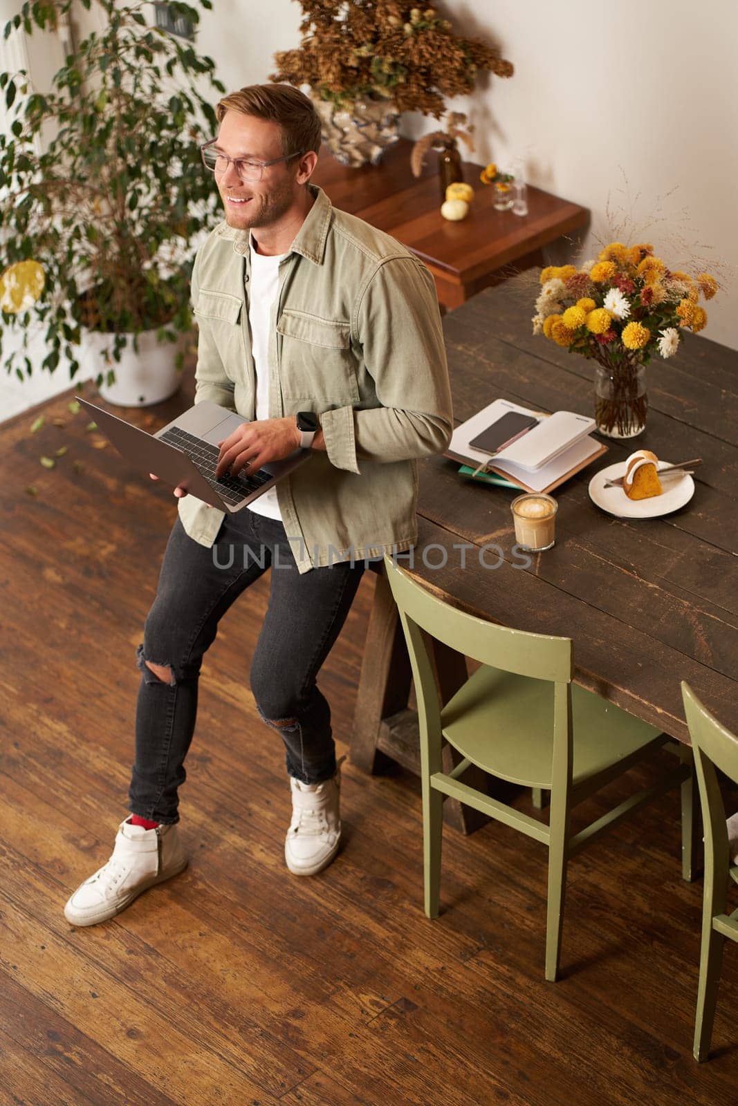 Vertical shot of handsome stylish businessman, standing near the table with laptop, thinking, working on a project on his computer, studying online, doing freelance task.