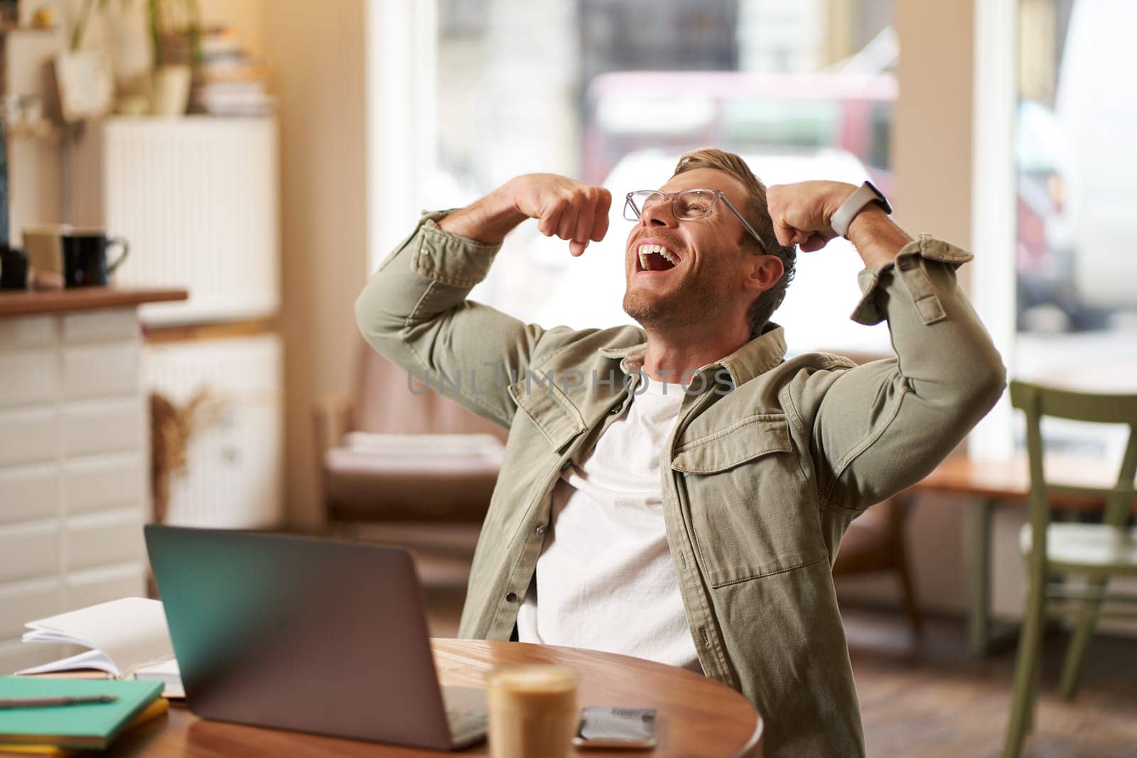 Portrait of pleased, smiling young man shouting with rejoice, sitting in cafe in front of laptop, flexing muscles, screaming from happiness, winning and celebrating.