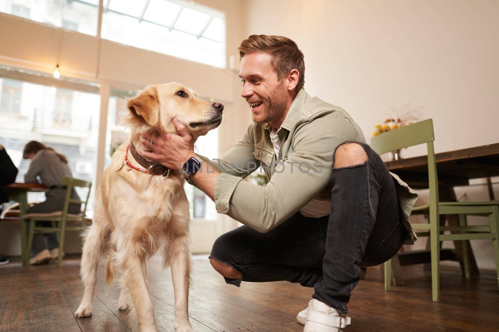 Portrait of happy young man enters coffee shop with his golden retriever. Handsome guy petting dog in pet-friendly cafe.