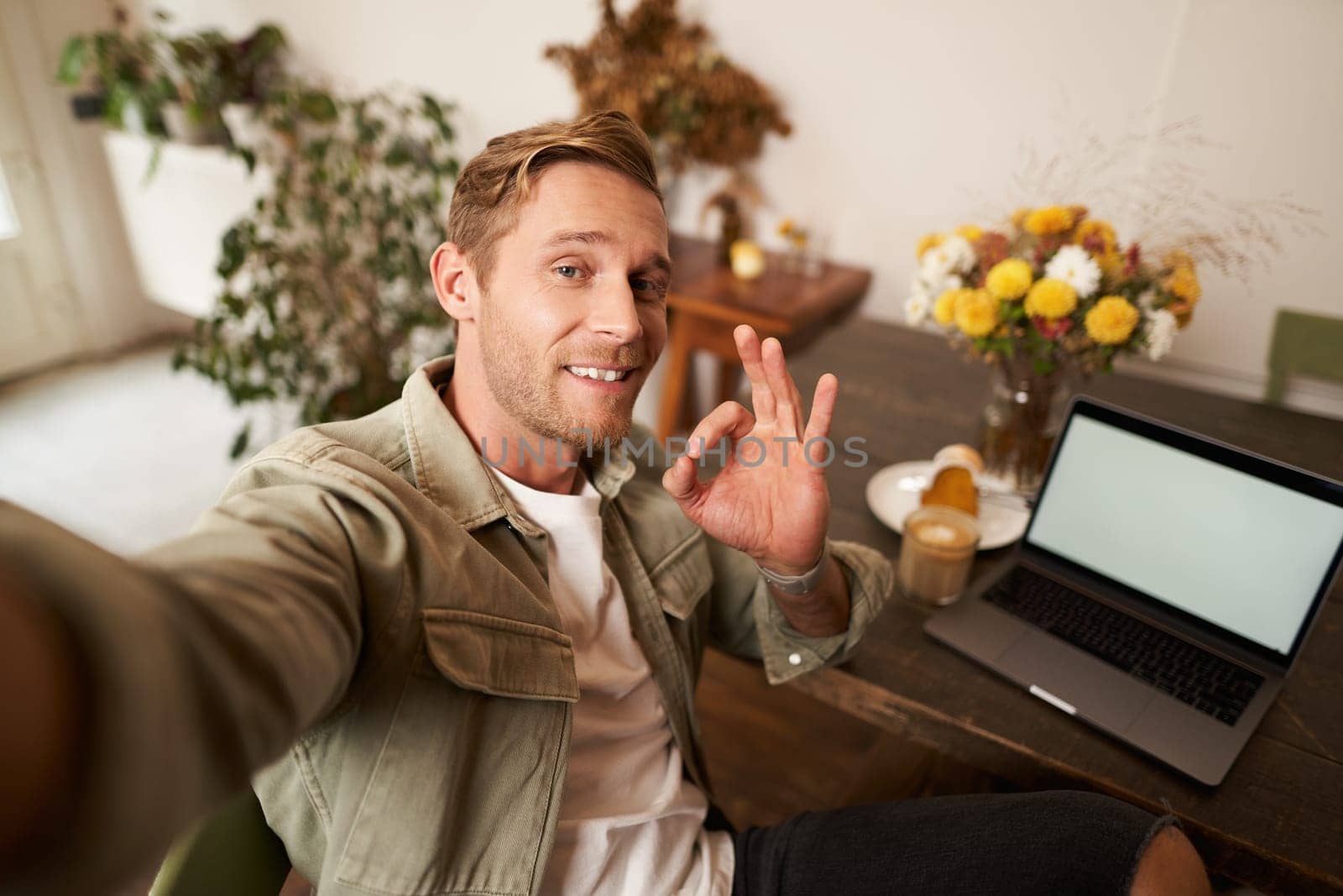 Portrait of young handsome man, cafe visitor taking selfie on smartphone app, showing okay, good recommendation sign, blogger rates coffee shop, nods in approval by Benzoix