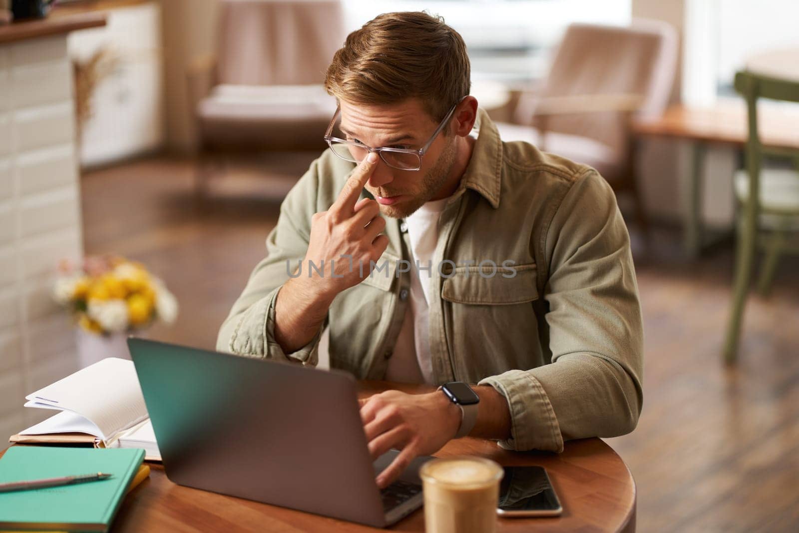 Image of young digital nomad, man in glasses sits in cafe, works from coffee shop, uses laptop in co-working space, wears glasses, drinks his beverage.