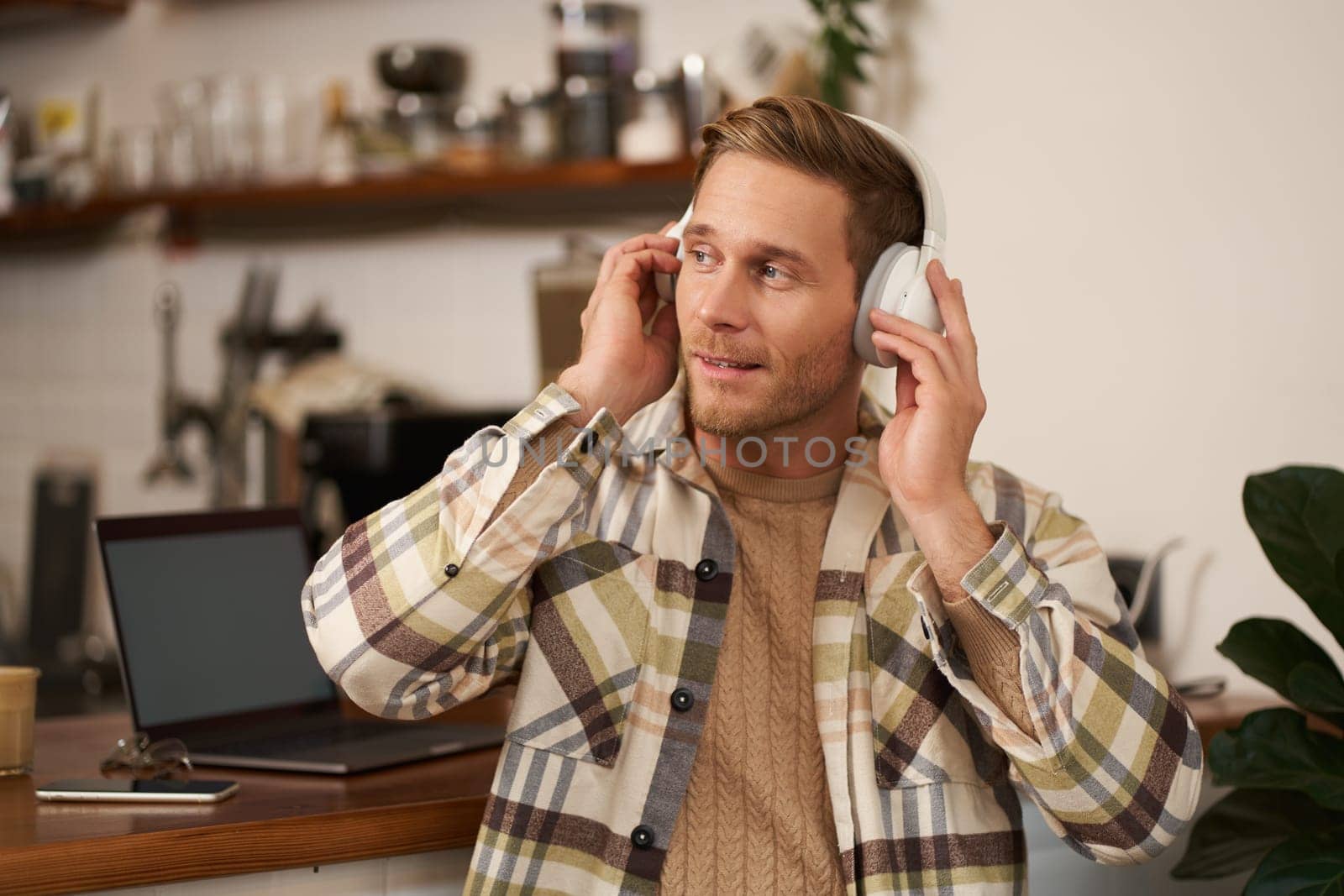 Lifestyle portrait of happy, excited young man, sitting in coffee shop with laptop and headphones, dancing on his chair and enjoying great quality sound, streaming favourite songs.