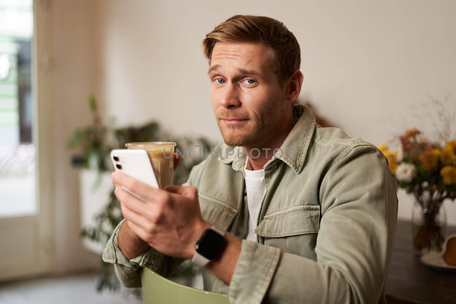 Portrait of young man in cafe, holding cup of coffee and mobile phone, looking at camera with skeptical face and a smirk by Benzoix