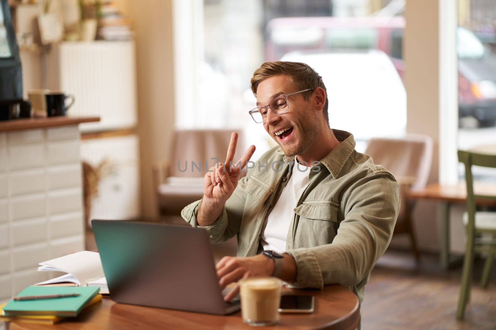Happy smiling guy in glasses, sits in cafe, shows peace sign at laptop camera, video chats, connects to online meeting.