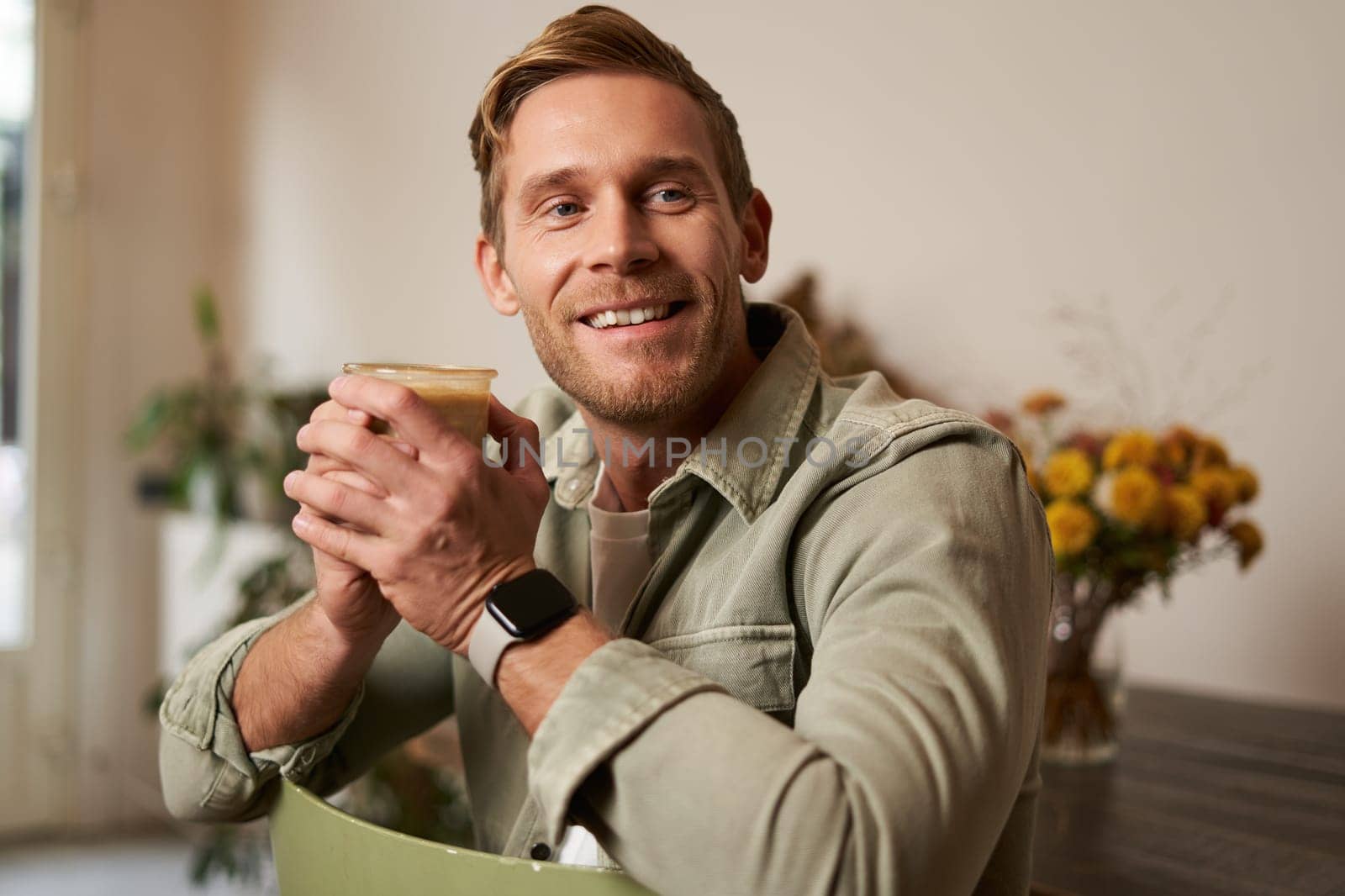 Portrait of good-looking young man with cup of coffee, sitting on chair in cafe, smiling and relaxing with his cappuccino drink.