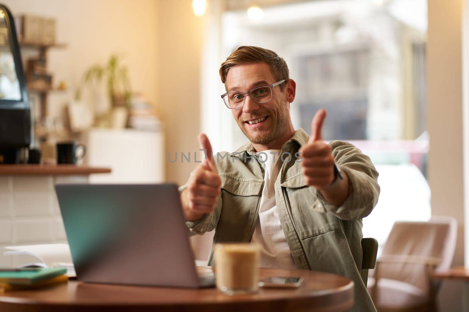 Joyful smiling guy in glasses, man shows thumbs up, sits in cafe with laptop, recommends website, e-learning service or co-working space.