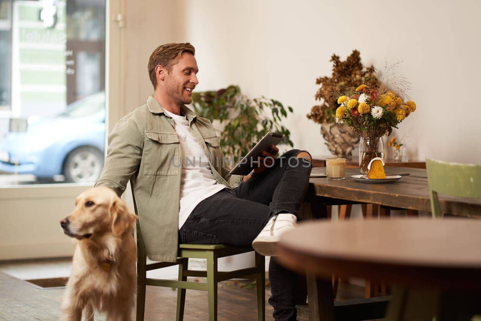 Portrait of handsome young man, cafe visitor, sitting with his dog, petting golden retriever and reading news on digital tablet, concept of lifestyle and pet-friendly public places by Benzoix