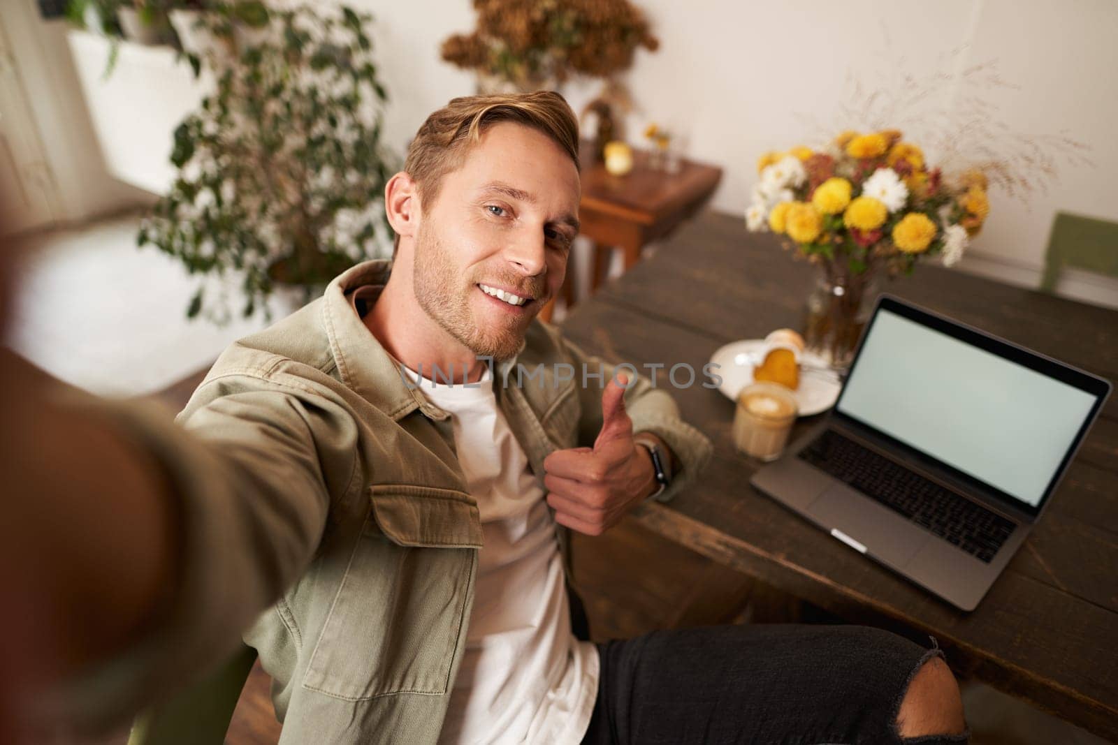 Man taking selfie with thumbs up, sitting in cafe with his laptop and smiling at camera, blogger posting on social media app.