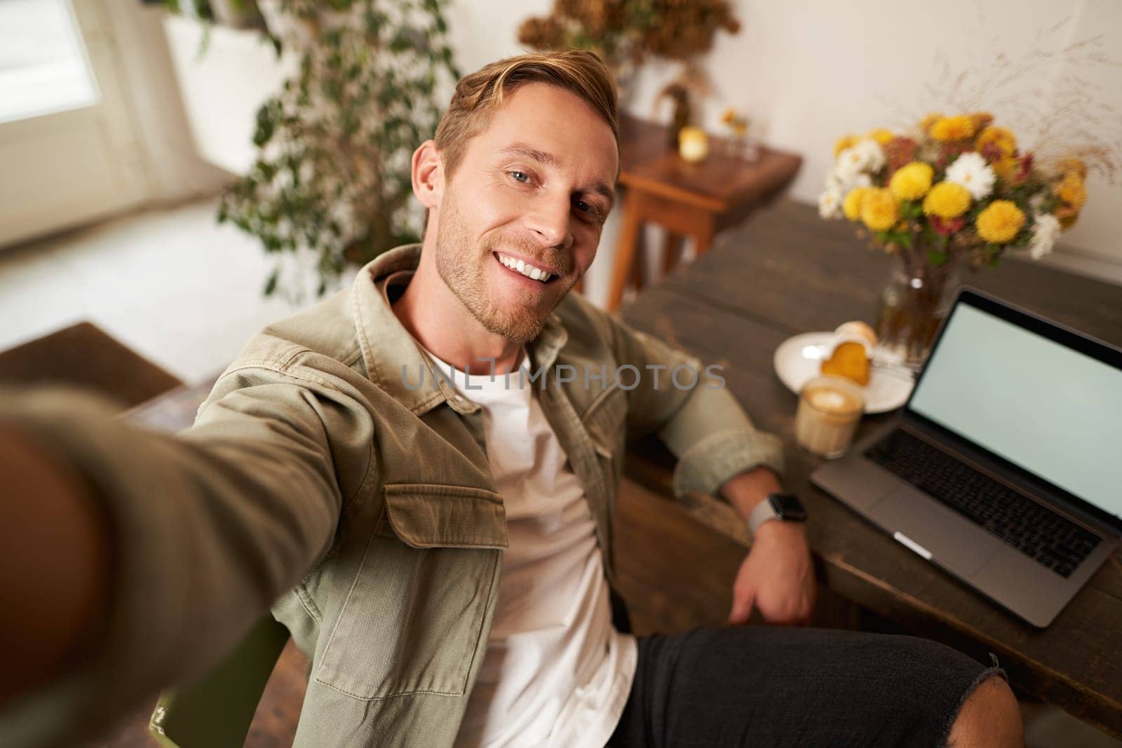 Selfie portrait of handsome young man, taking picture of himself in cafe with laptop, smiling at camera.