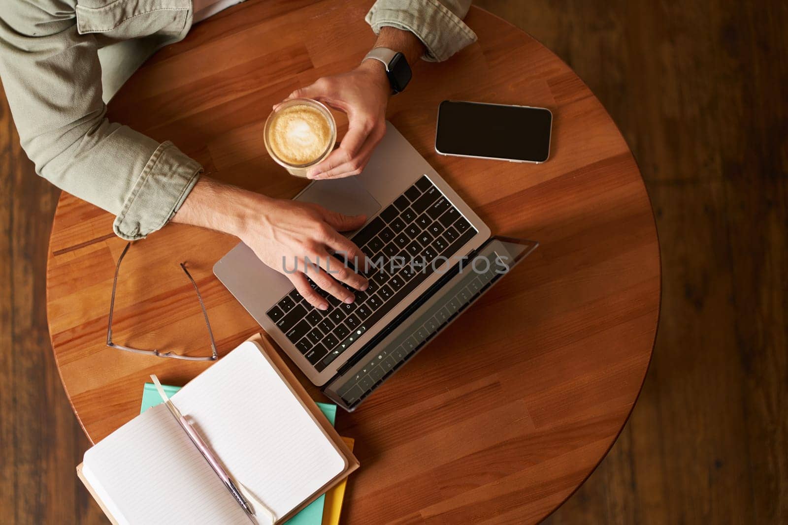 Co-working space, freelance and e-learning concept. Top view, male hands holding cup of coffee and using laptop. A man sitting in cafe and working or studying online.