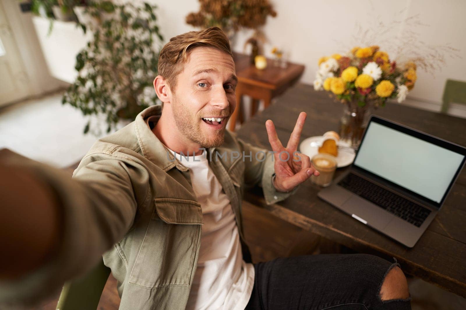 Selfie portrait of handsome young man, taking picture of himself in cafe with laptop, smiling at camera by Benzoix