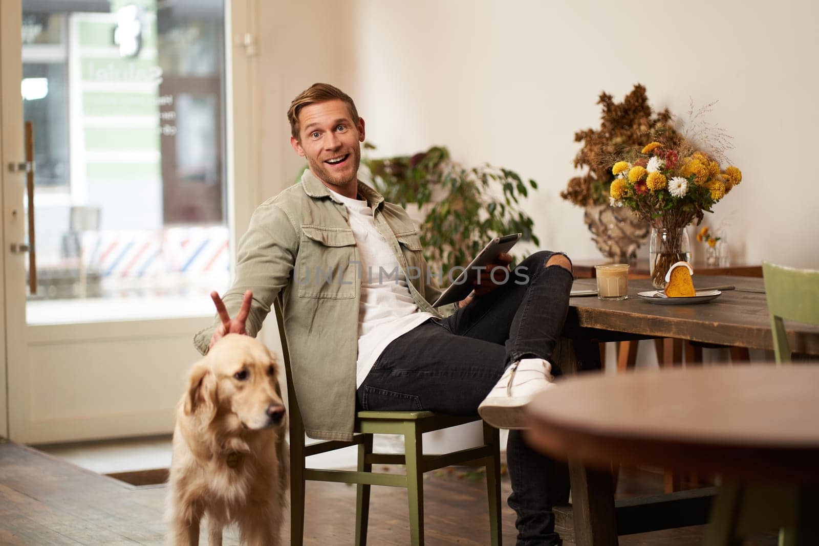 Portrait of happy smiling young man, cafe visitor, sitting in coffee shop with his dog, petting golden retriever, holding digital tablet, reading news or browsing on internet by Benzoix