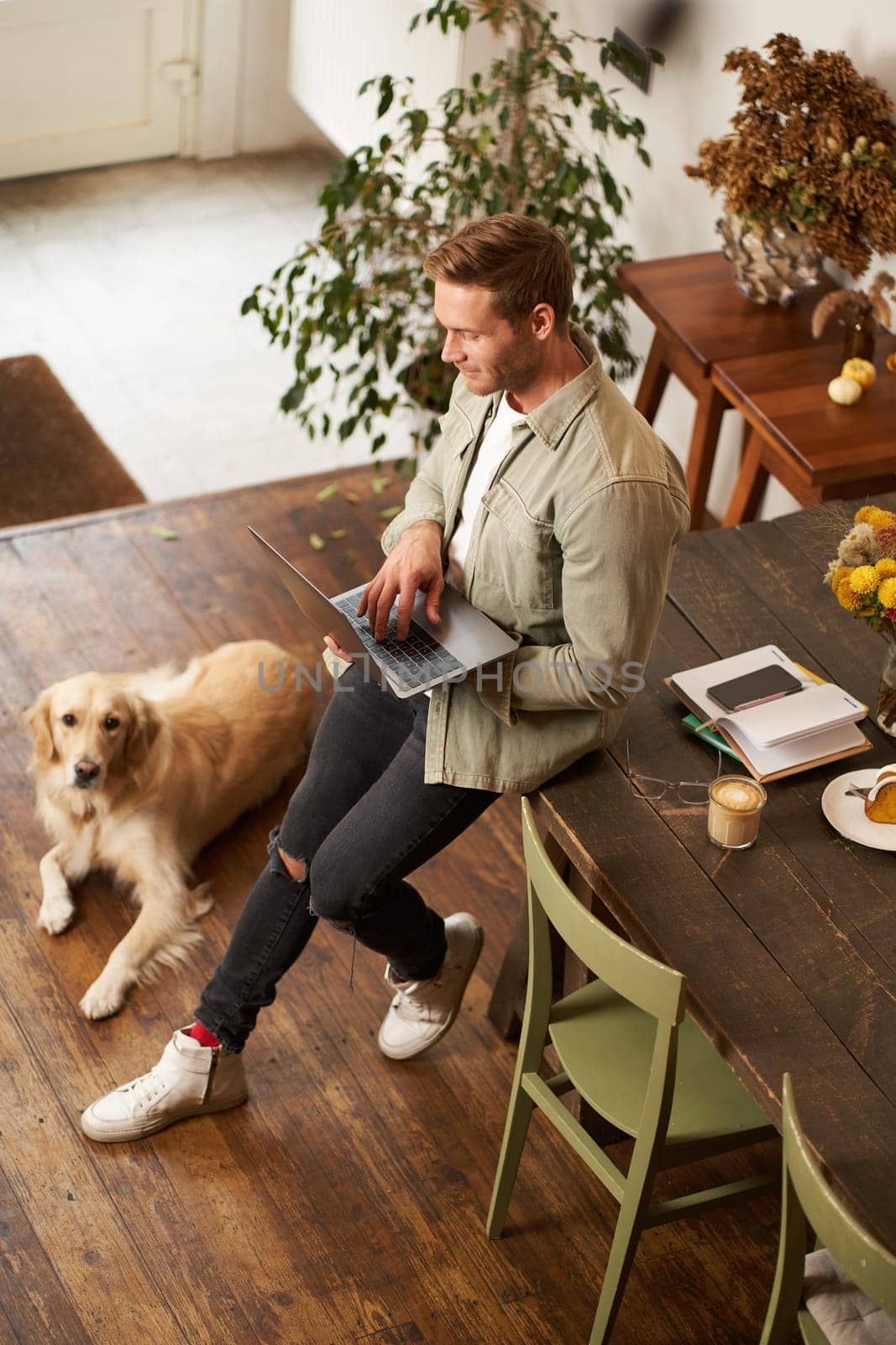 Vertical shot of handsome young man with a dog, leaning on table, holding laptop in hands, looking at screen, checking emails at work. Businessman at co-working space with his golden retriever.