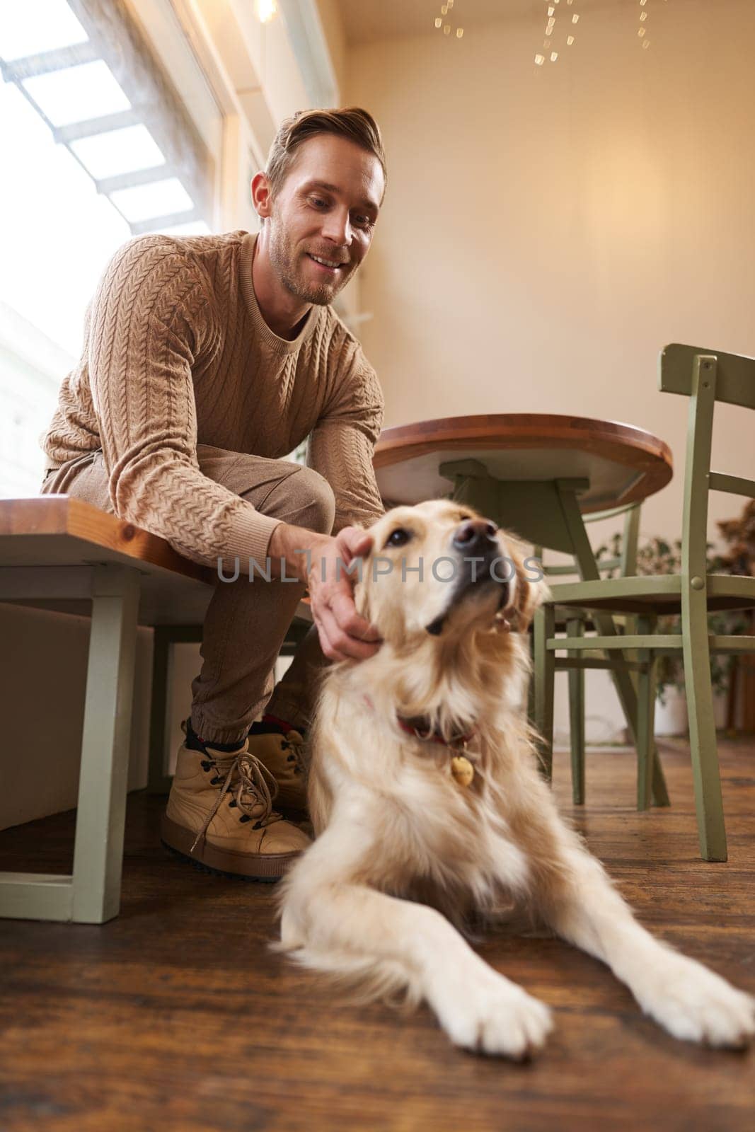 Vertical shot of young smiling man with his dog, golden retriever pet in a cafe.