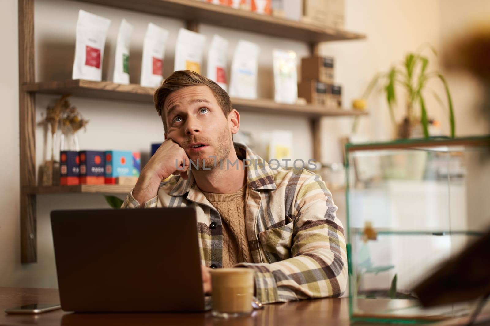 Portrait of stressed working man, sitting in cafe with laptop, business owner doing project online, looking up, roll his eyes, drinking coffee in co-working creative space.