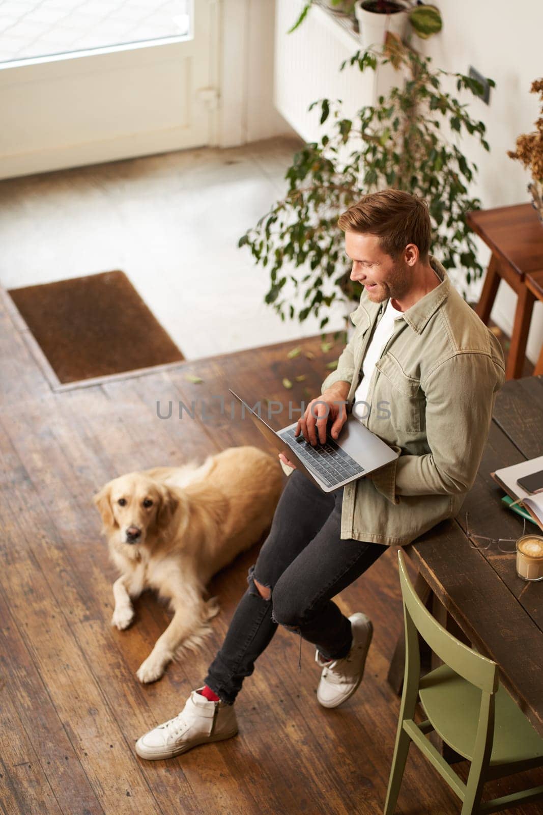 Vertical shot of handsome businessman, young man with laptop leans on table, sitting at co-working space with his dog. Young salesman checks his messages, smiling, working in an office with a pet.