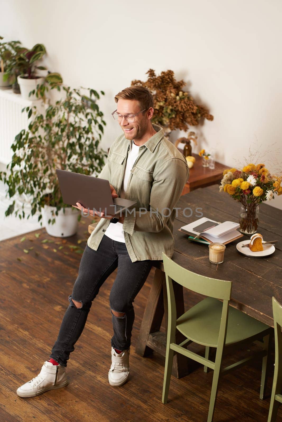 Vertical portrait of handsome happy man, manager or CEO of company, sitting on table and holding laptop, laughing, video chatting, has an online meeting with a team, standing in an office by Benzoix