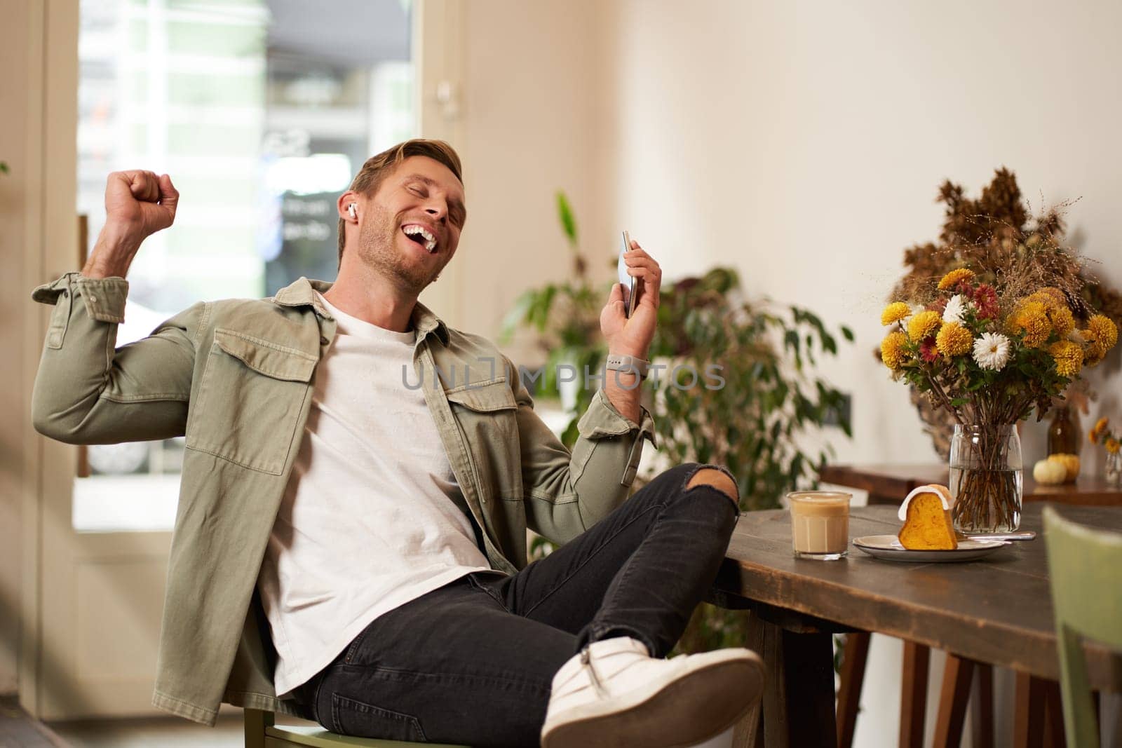 Portrait of handsome happy man in cafe, listens to music in wireless earphones, holding smartphone, connects to public wifi and enjoys favourite song, relaxing in coffee shop.